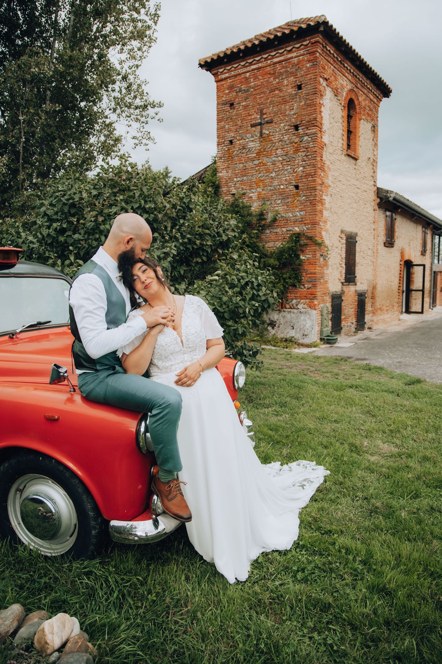 Bride and groom embrace leaning on vintage red car beside rustic brick tower.