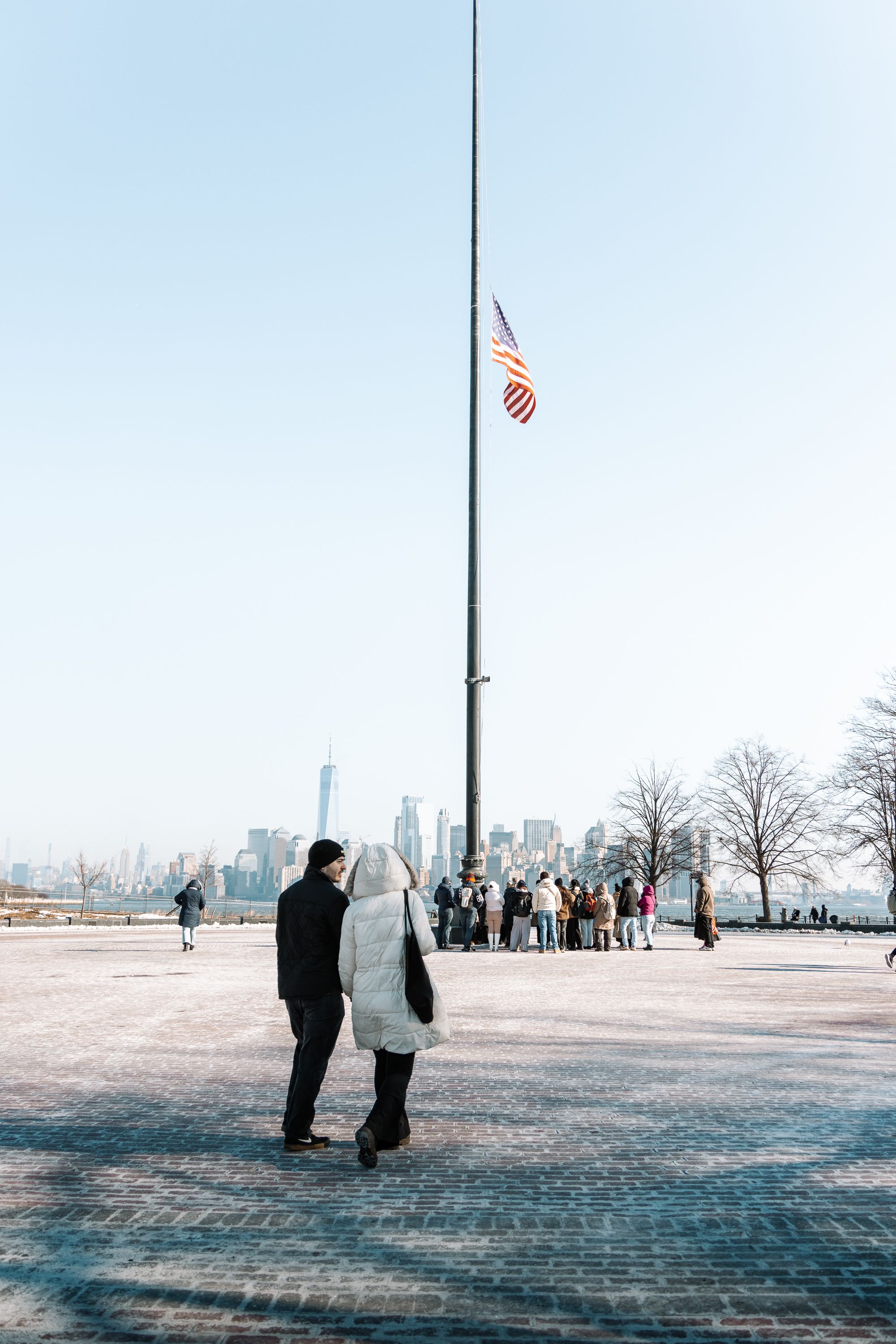 People walking on a snowy plaza with the New York City skyline visible in the distance.