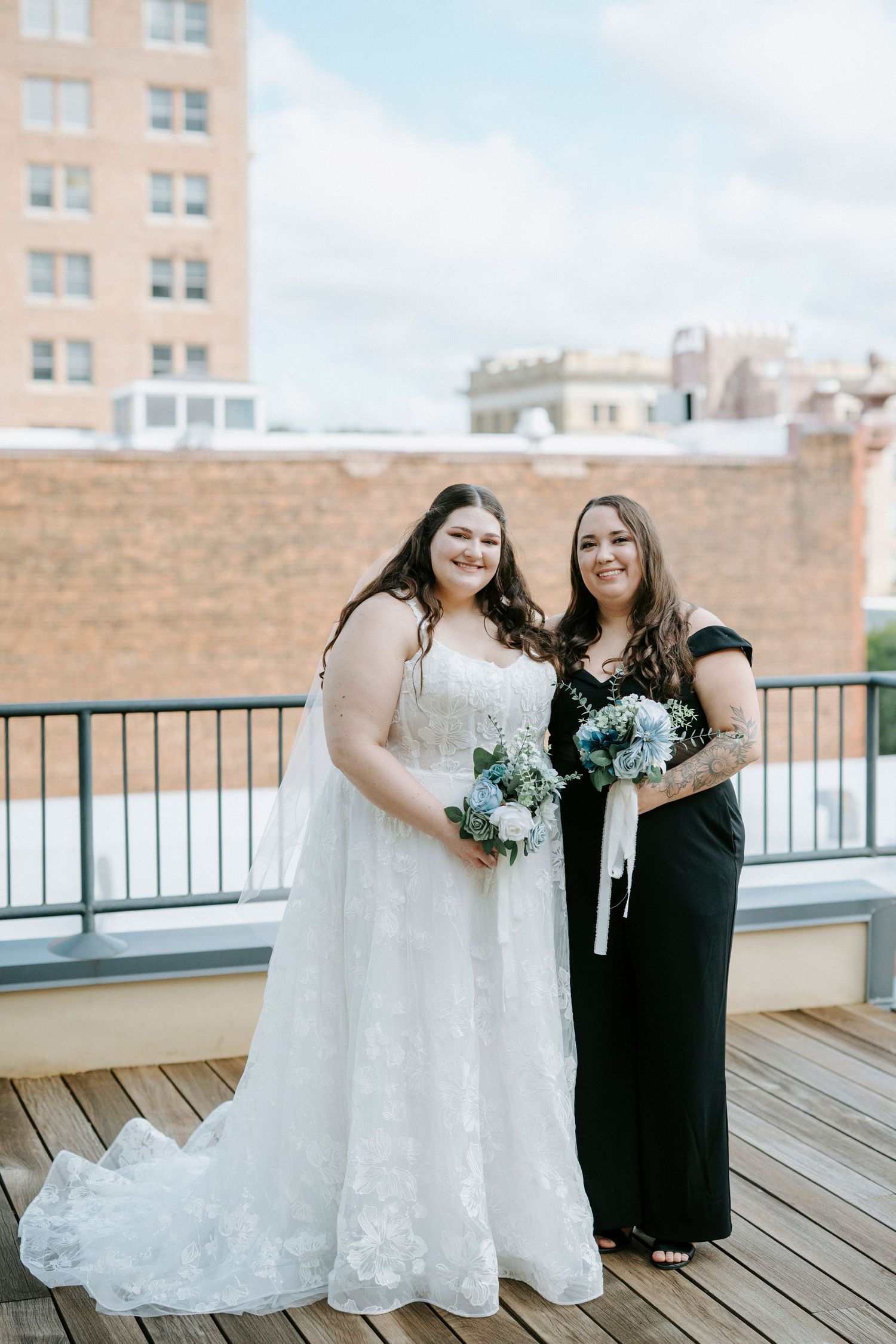 Two people in contrasting black and white dresses stand together on a wooden deck with buildings in the background.