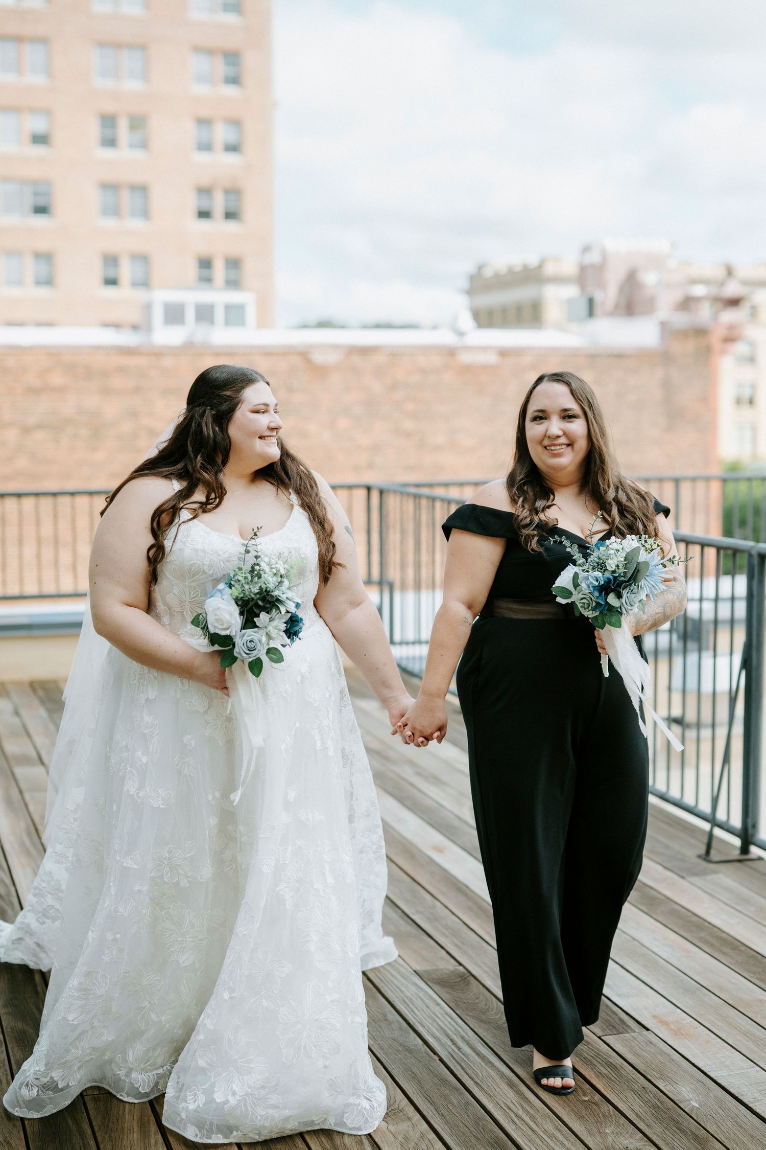 Two people share a joyful moment on a rooftop deck with one wearing a white wedding gown and the other in black.