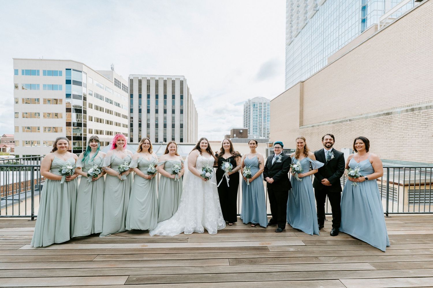 Wedding party in blue dresses poses on a rooftop terrace with city buildings in the background.