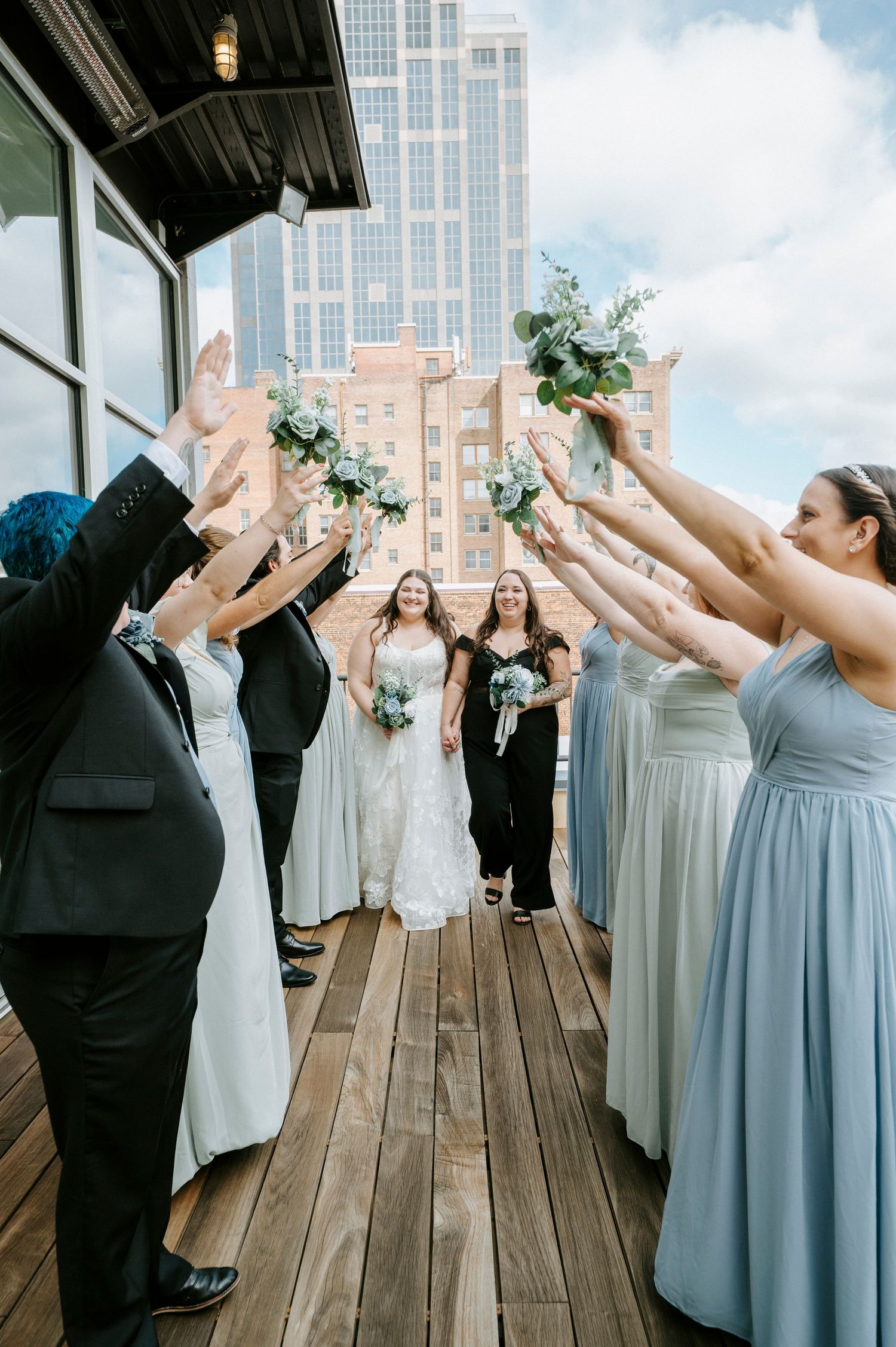 Wedding party celebrates on wooden deck with bridesmaids in light blue dresses and groomsmen in dark suits.