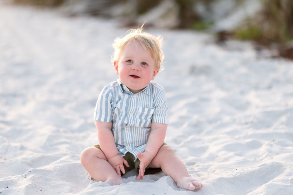 Baby sitting on pristine white sand beach wearing blue and white striped outfit.