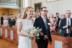 Bruden og hennes far nesten i enden av kirkegulvet. De smiler og bruden holder en brudebukett.
