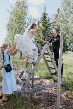 Brud i joggesko klatrende over et gjerde. Brudgom står for å ta henne i mot og forlovere løfter kjolen.
