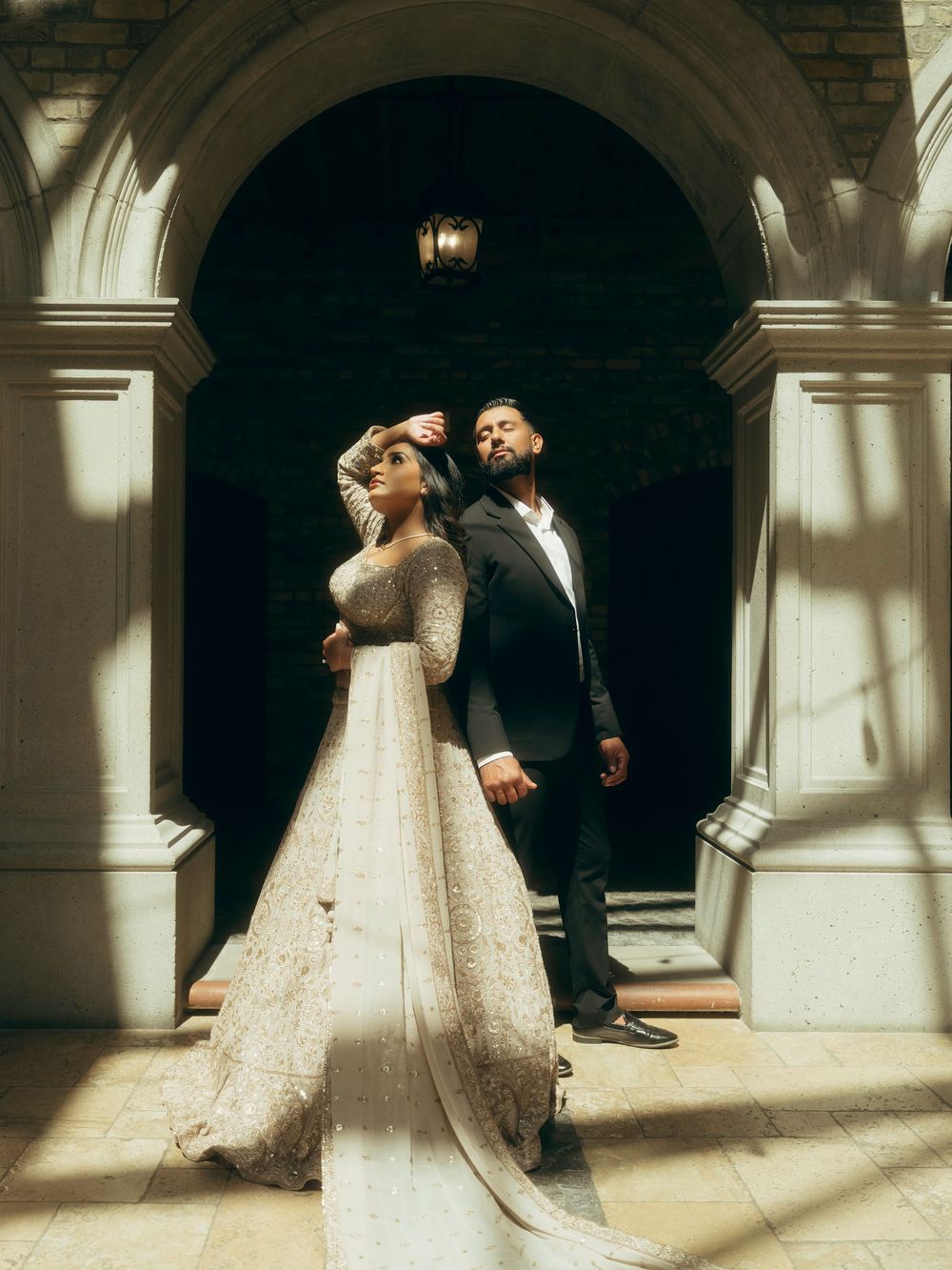 A romantic wedding couple dances in an elegant archway with dramatic lighting and shadows creating an intimate atmosphere.