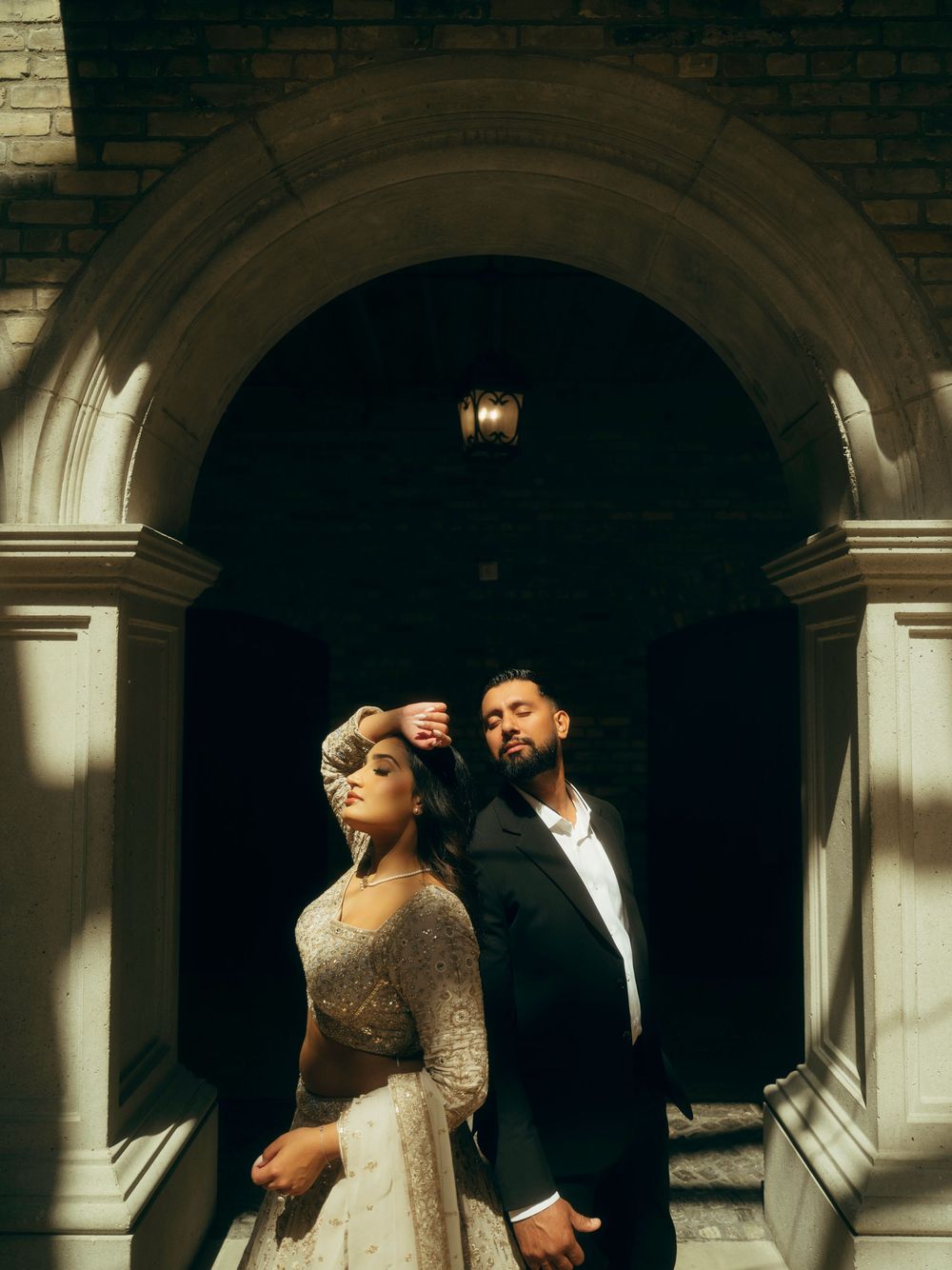 Romantic couple in formal attire embraces under arched stone colonnade in dramatic lighting.