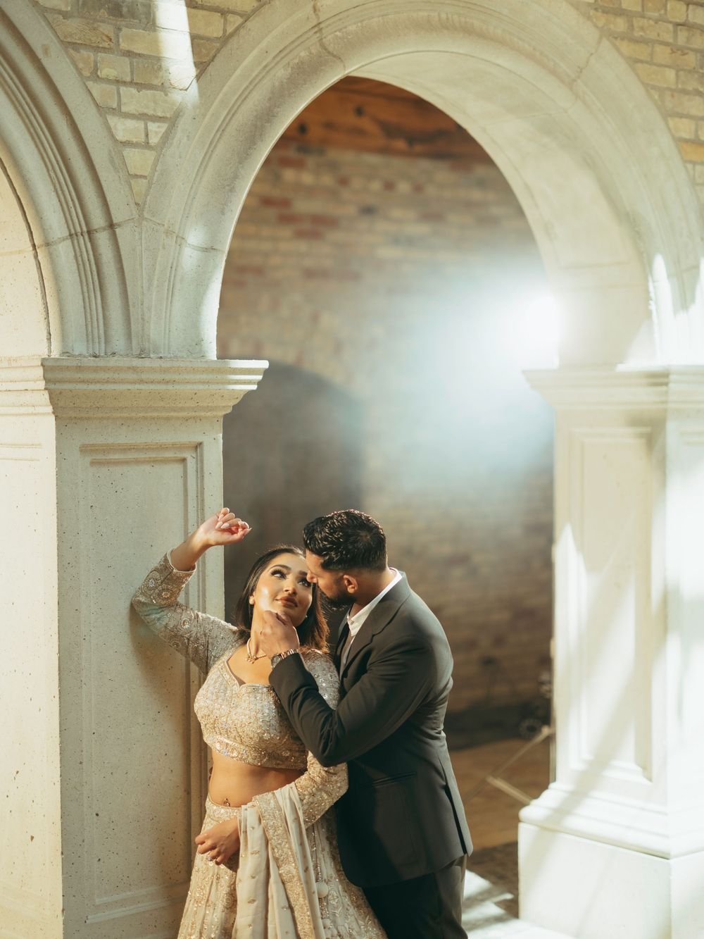 Romantic couple in traditional South Asian wedding attire embrace under stone archways with soft lighting.