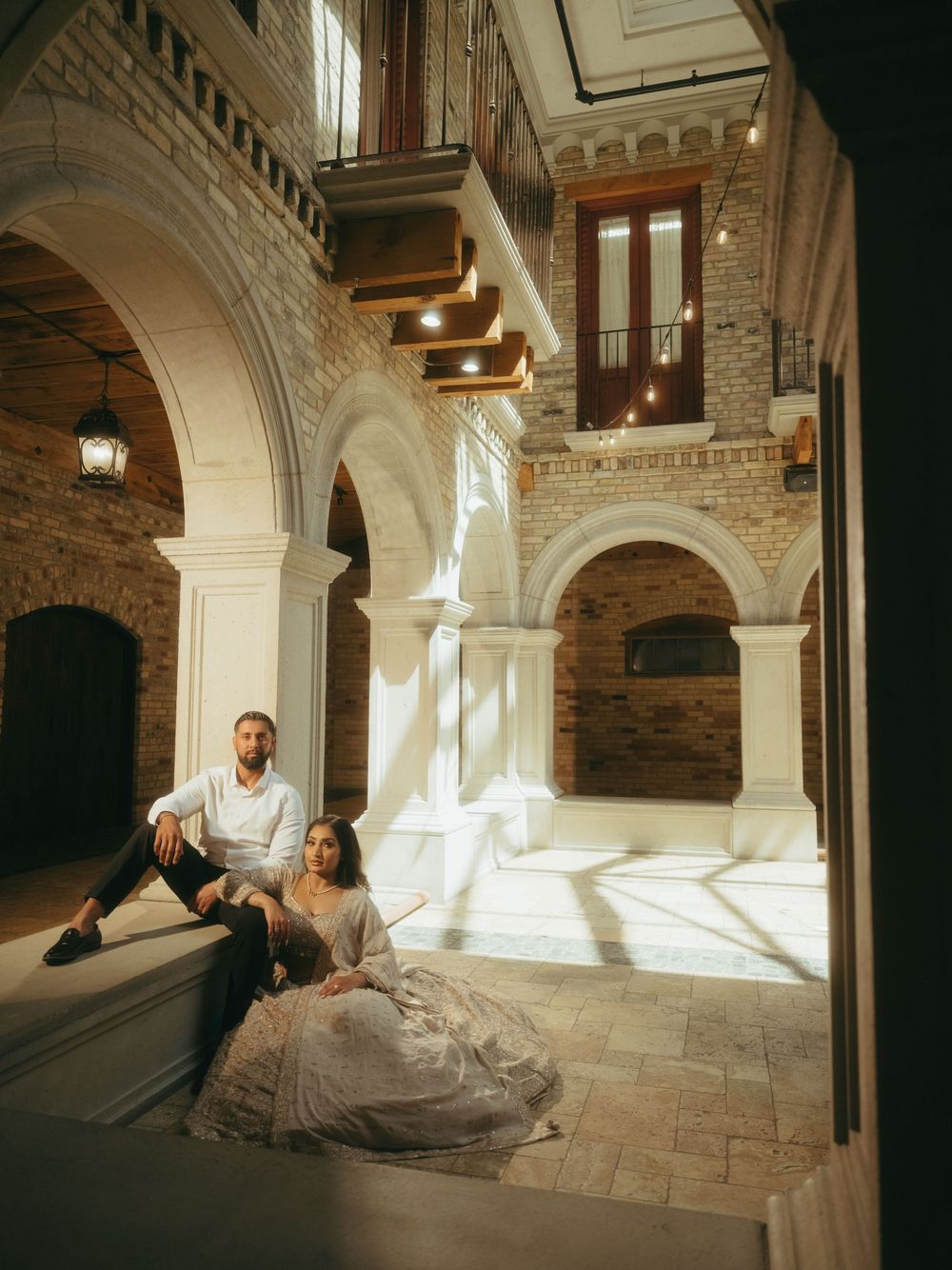 Elegant couple sits on stone steps inside historic building with arched architecture and natural sunlight streaming in.