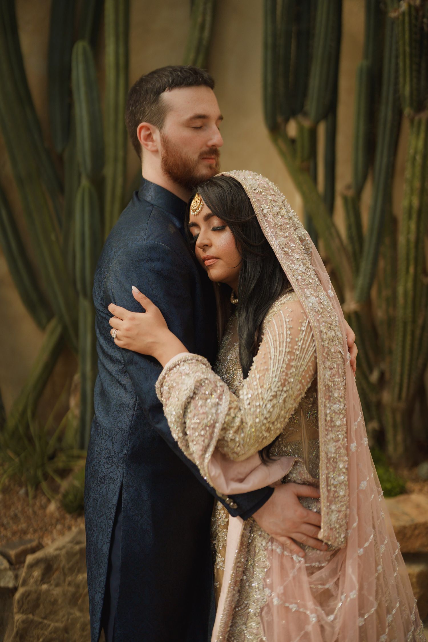 A romantic couple embraces in front of tall cacti, with the bride wearing an ornate beige and gold traditional South Asian outfit.
