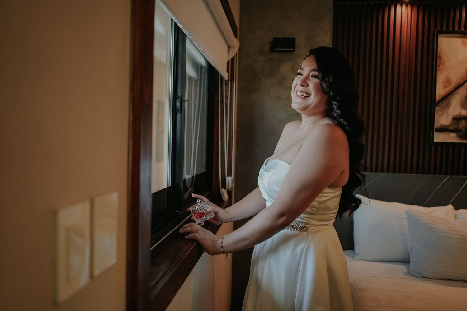 A bride in a white wedding dress stands in a warmly lit hotel room preparing for her special day.