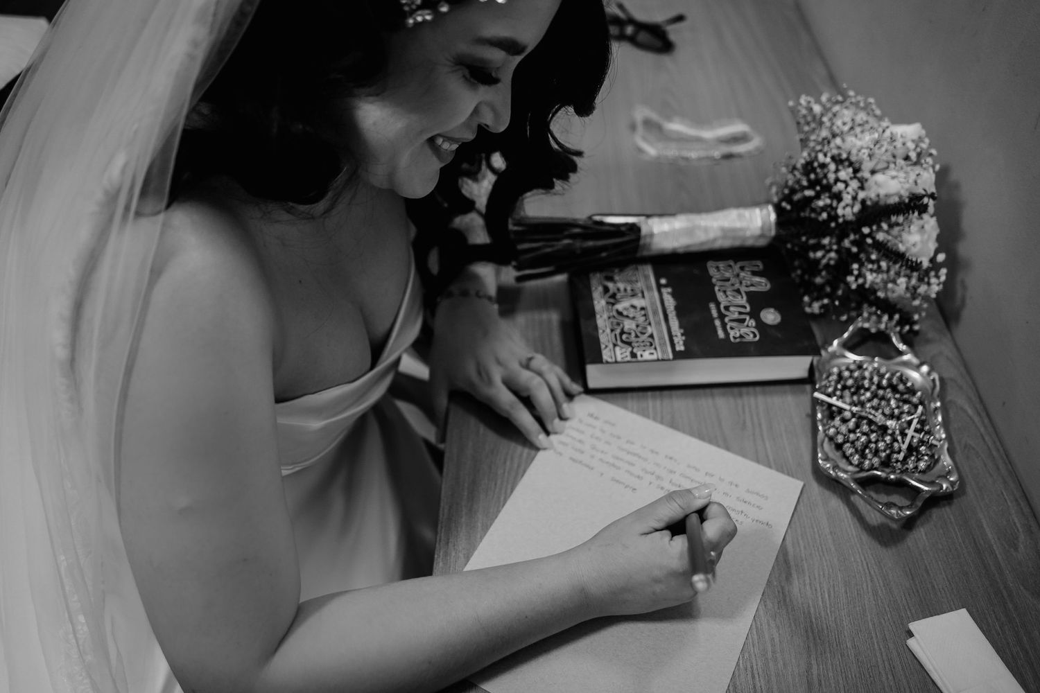 Black and white photo showing someone holding a camera while sitting at a desk with scattered papers and equipment.