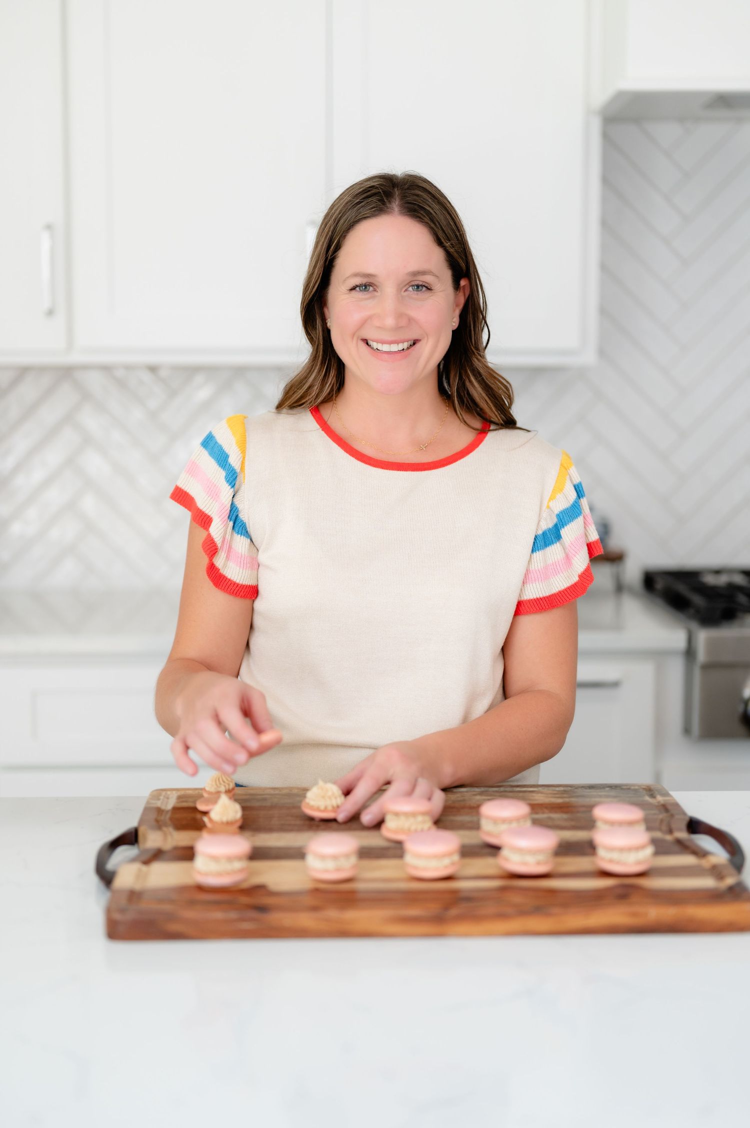 A person in a white top with colorful trim arranges pink macarons on a wooden serving board in a bright kitchen.