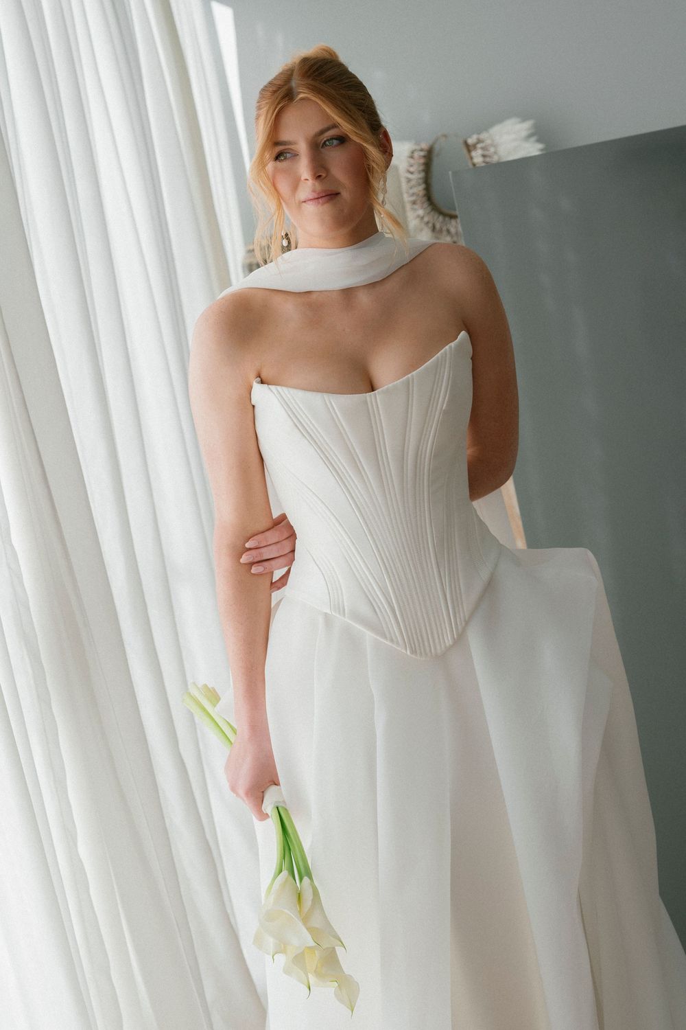 Elegant white structured wedding gown with architectural details photographed against sheer curtains.