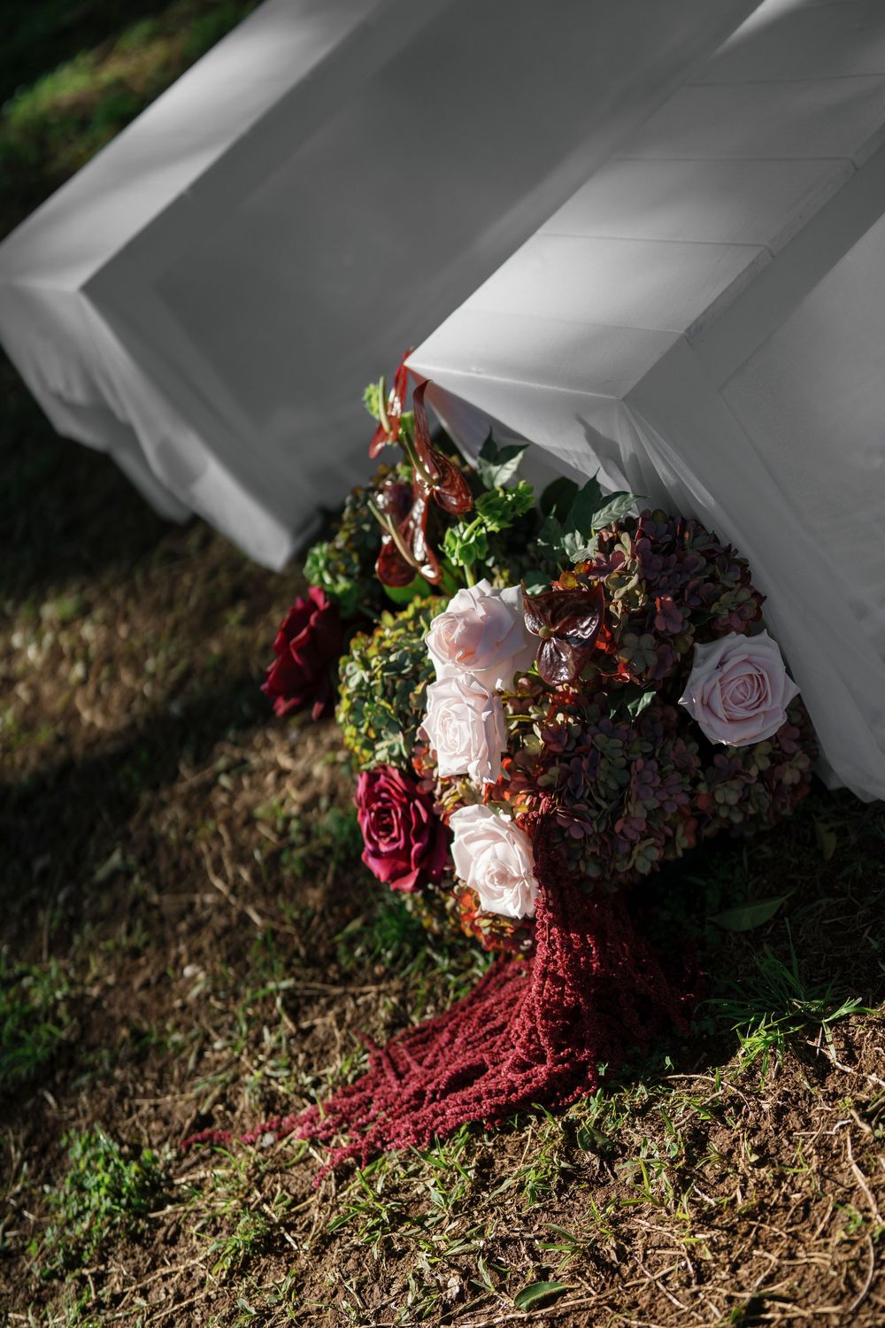 A bouquet with burgundy and pink flowers lies on grass near white fabric.