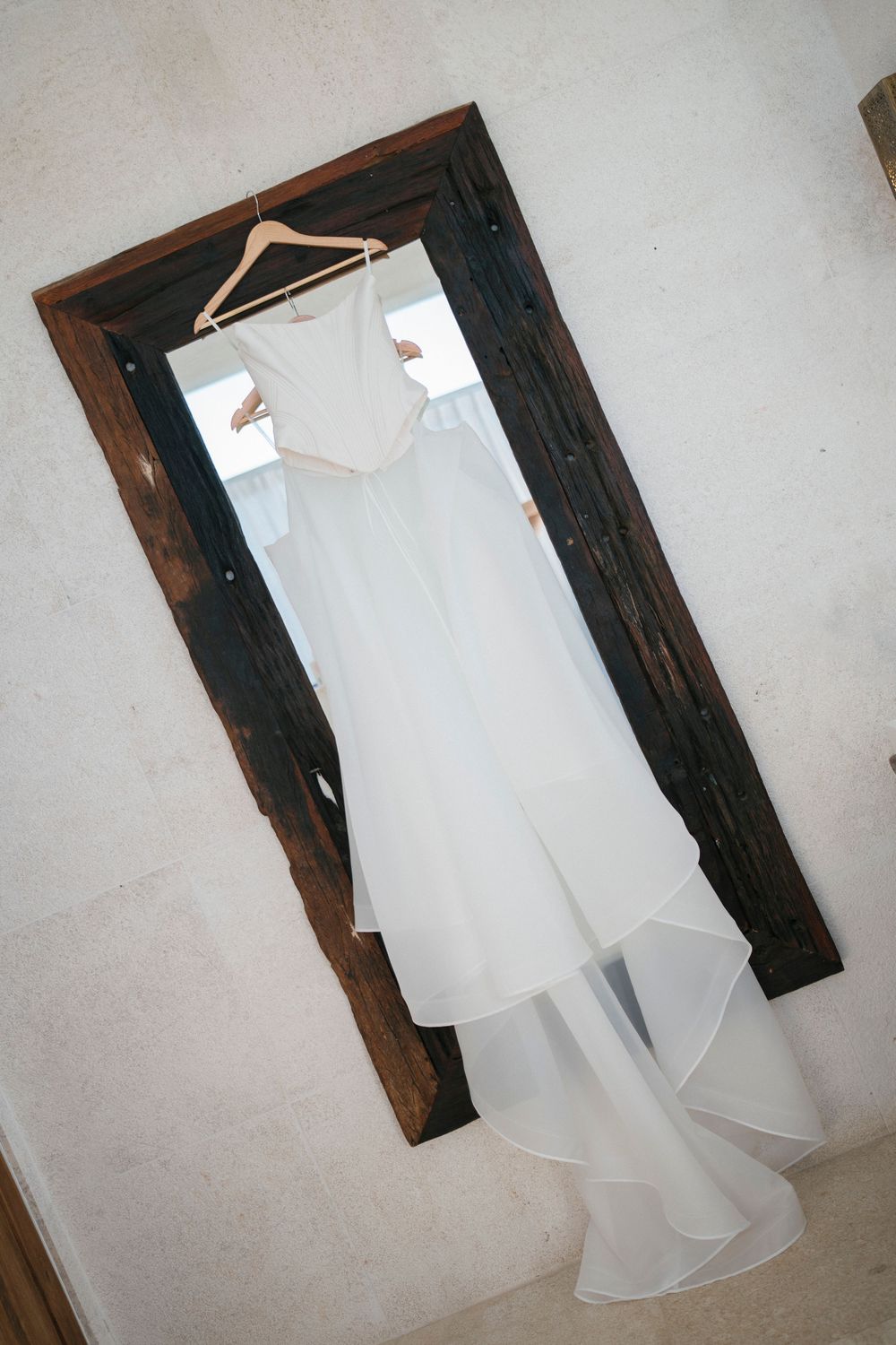 A white wedding dress hangs in front of a dark wooden mirror.