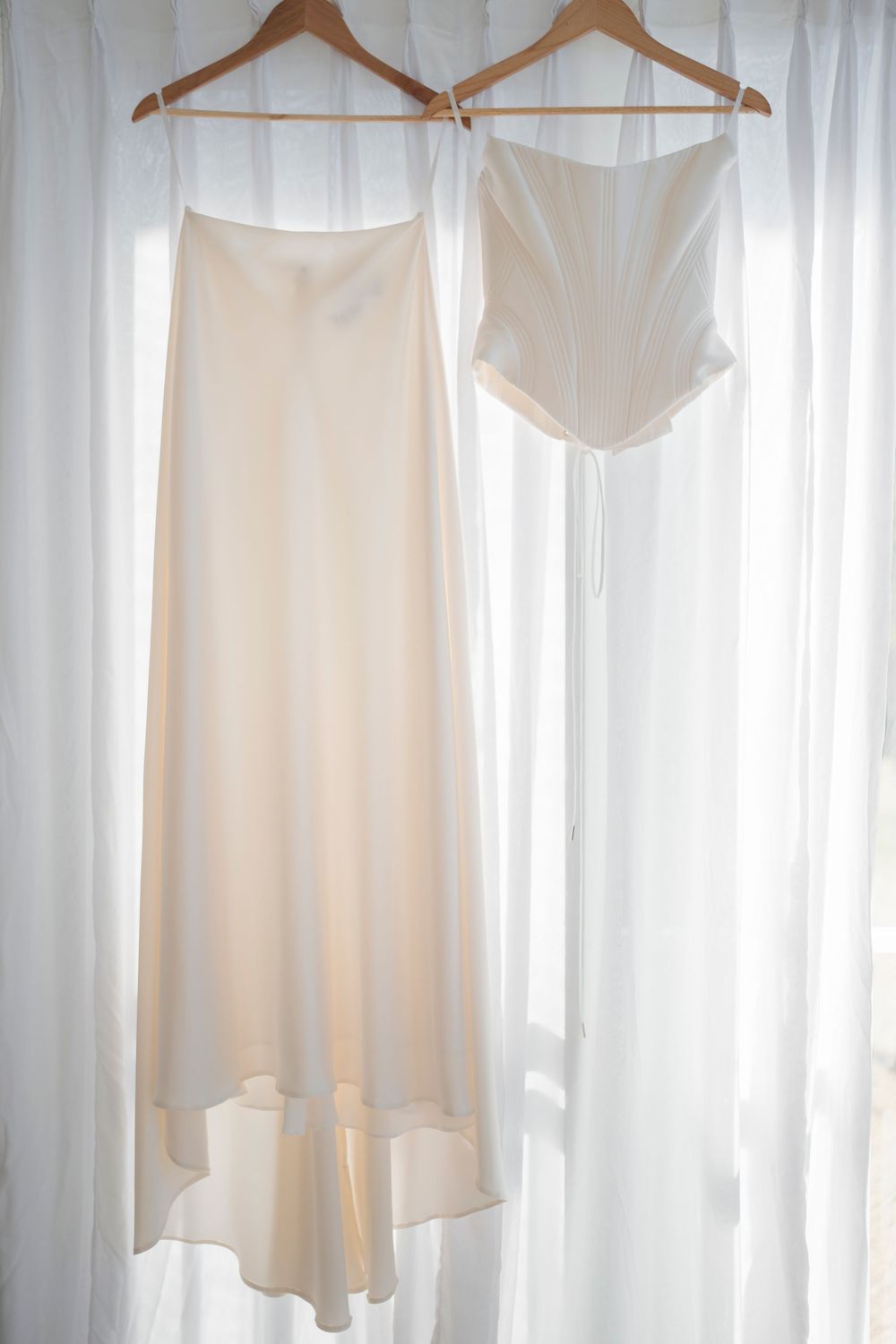 White wedding undergarments hanging on wooden hangers against sheer curtains.