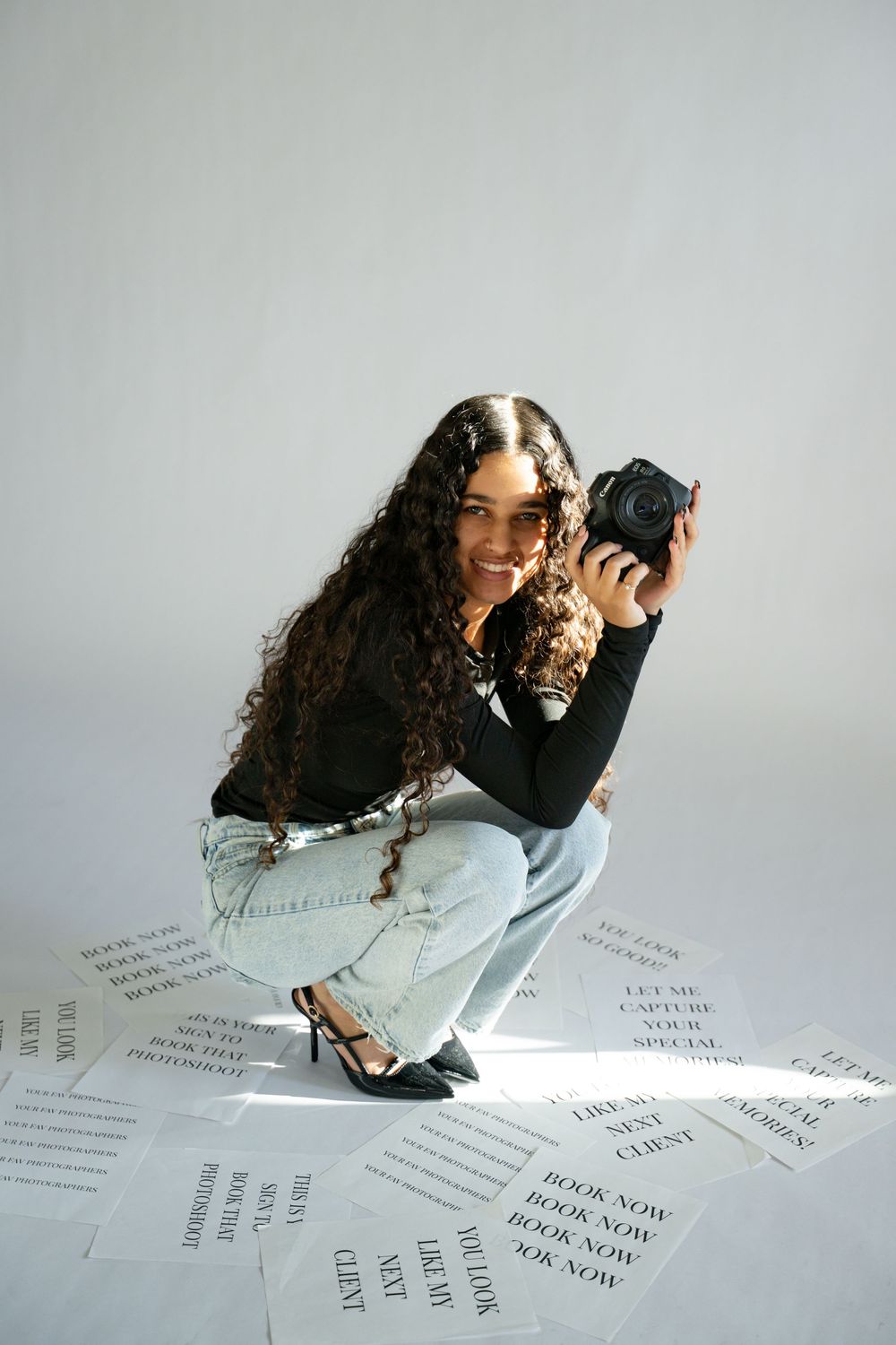 Photographer in black top and light jeans crouches on floor covered with paper text.