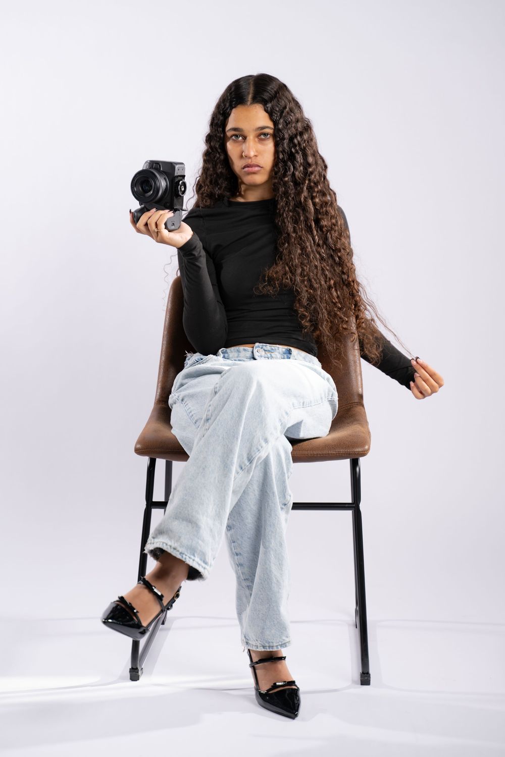 Photographer with long curly hair poses with camera while sitting on chair against white background.