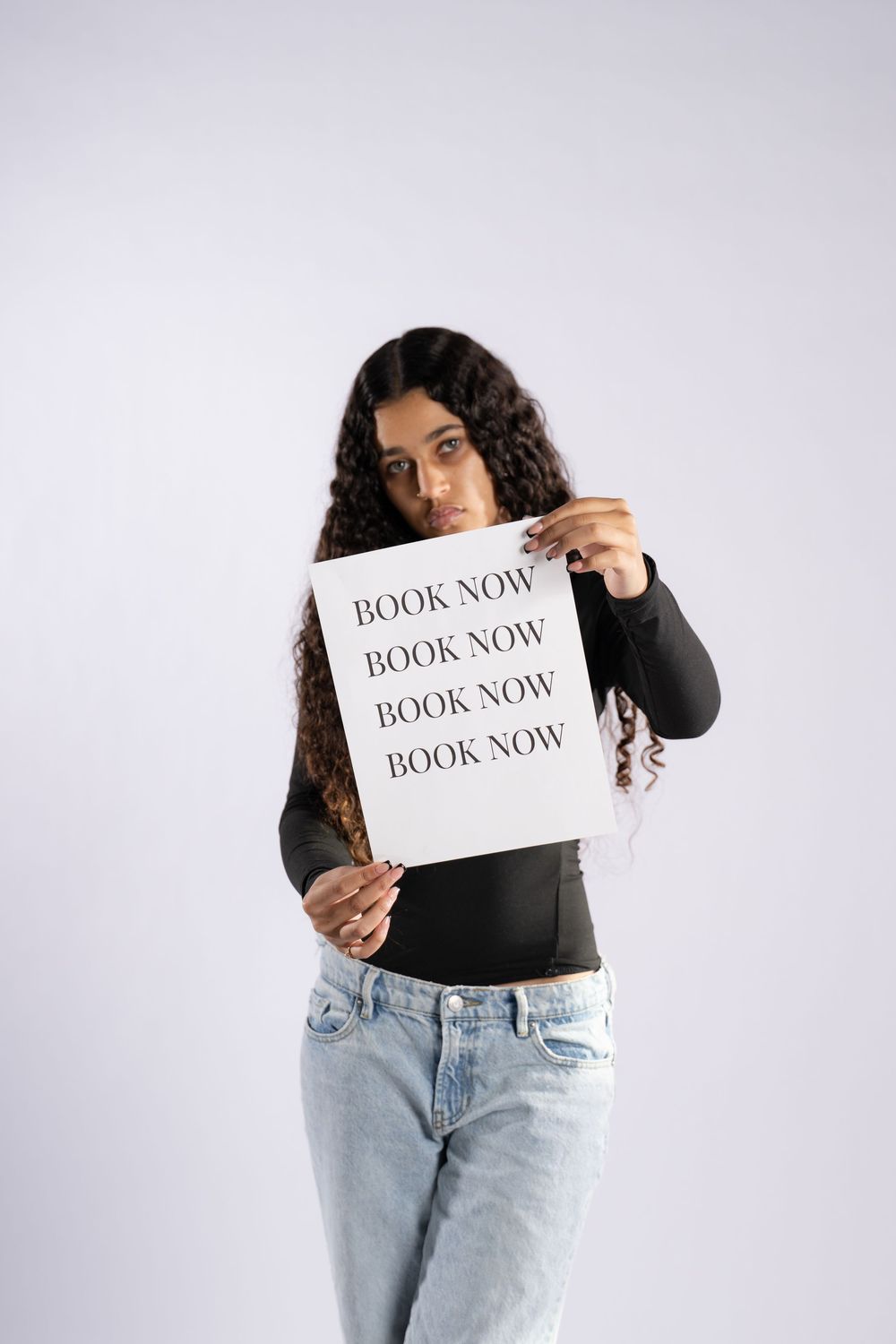 Model in casual wear holds white paper with 'book now' text repeated against light background.