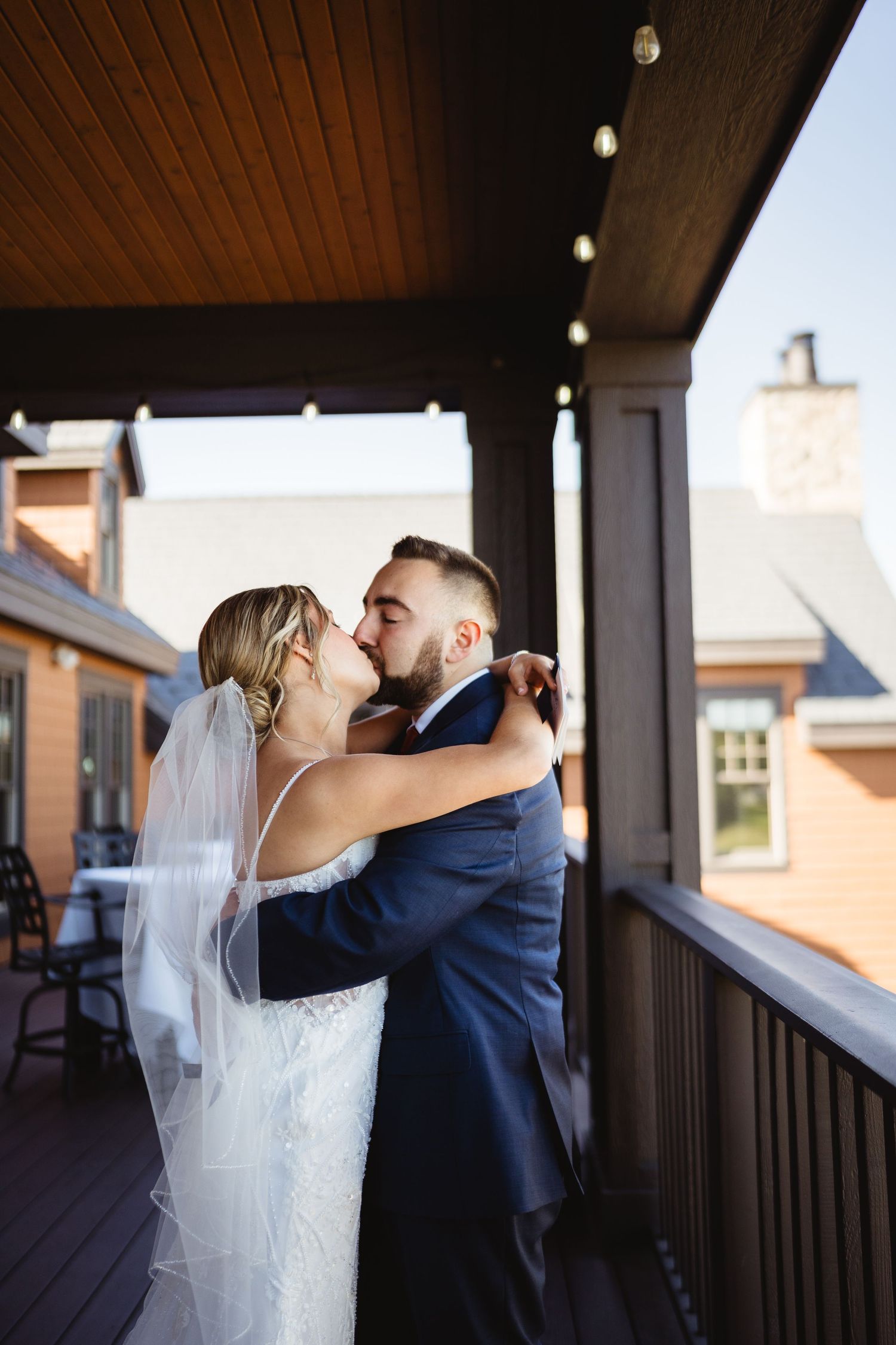 A couple in wedding attire share a romantic kiss on a covered porch with warm lighting and wooden beams overhead.
