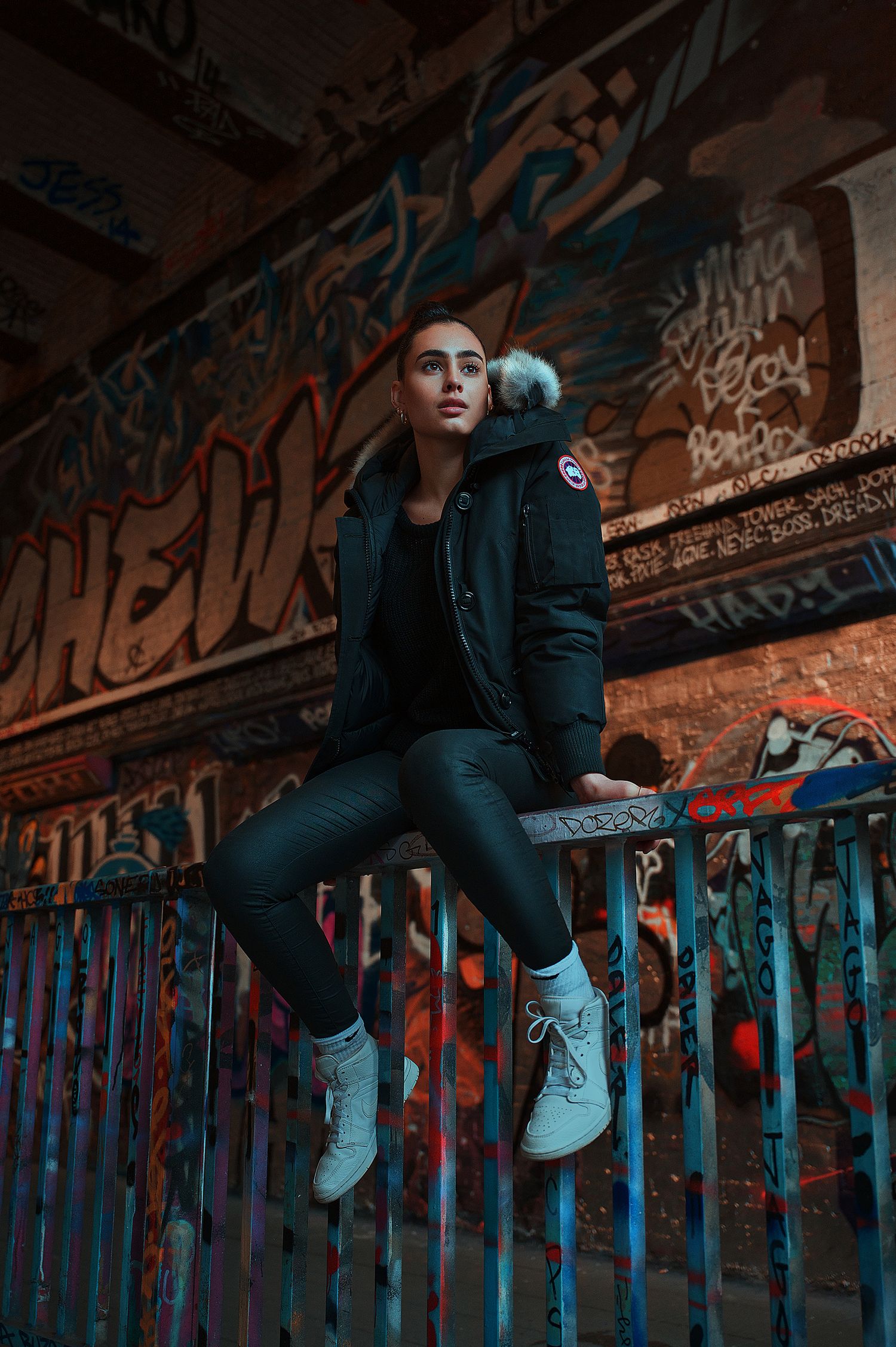 Person wearing black outfit and white sneakers sits on graffiti-covered ledge in moody urban setting with blue lighting.