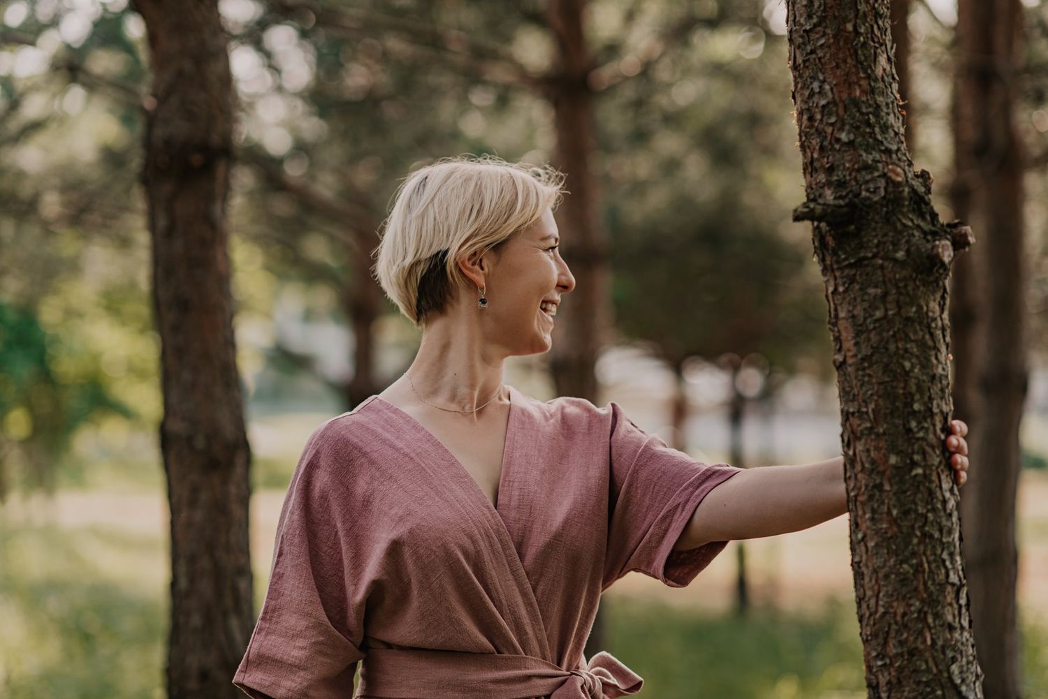 Someone in a dusty rose colored dress poses gracefully against a tree in a wooded setting.