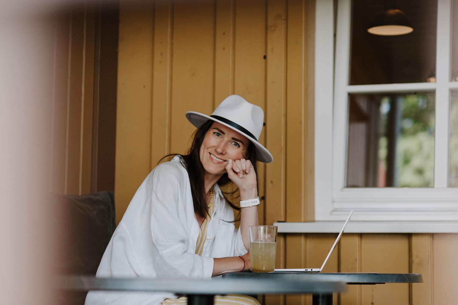 A woman in a gray hat sits at an outdoor table with a laptop, resting her head on her hand.