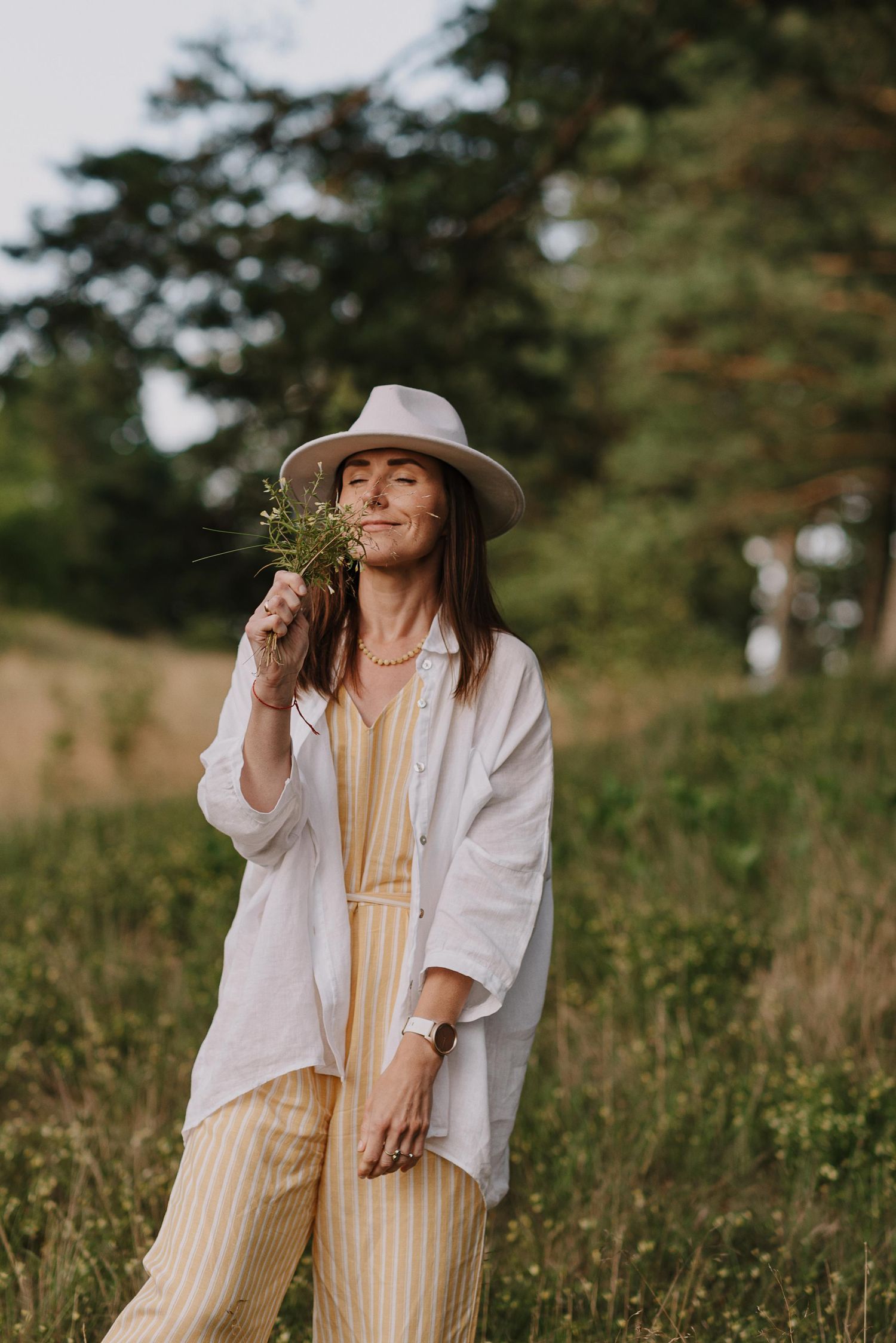 A woman in a white shirt and hat holds wildflowers in a sunny meadow.