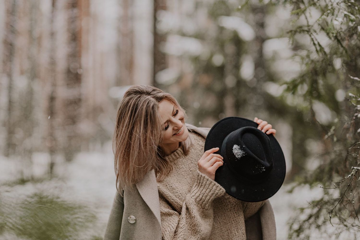 Stylish winter outfit featuring a black hat and beige sweater photographed in a snowy forest.