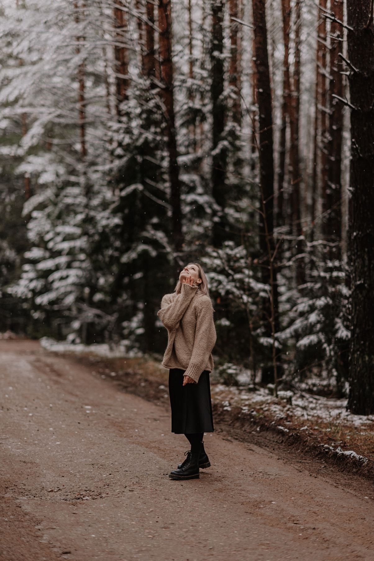 A figure in a beige coat stands on a snowy forest path surrounded by tall trees.