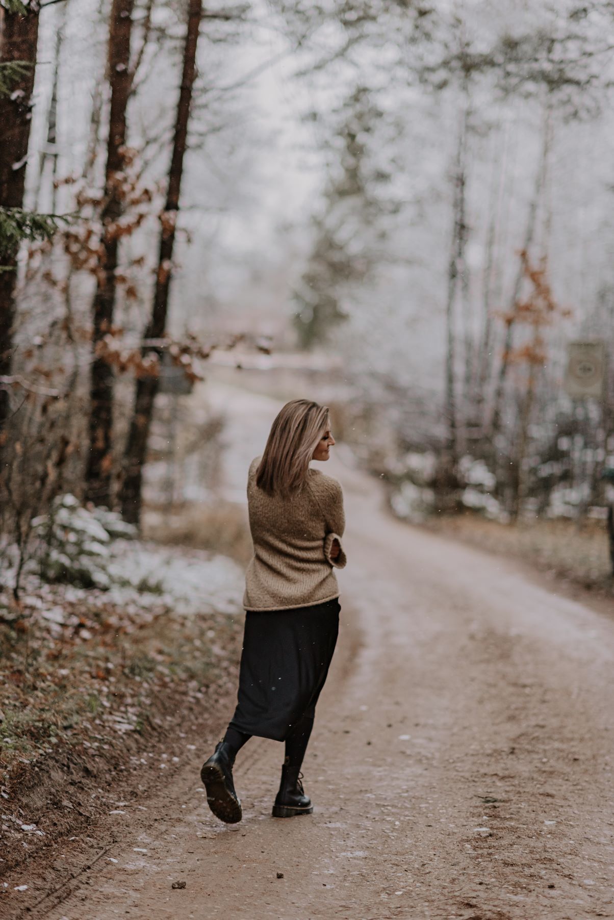 A figure in a beige top and dark pants walks along a winding forest trail in winter.