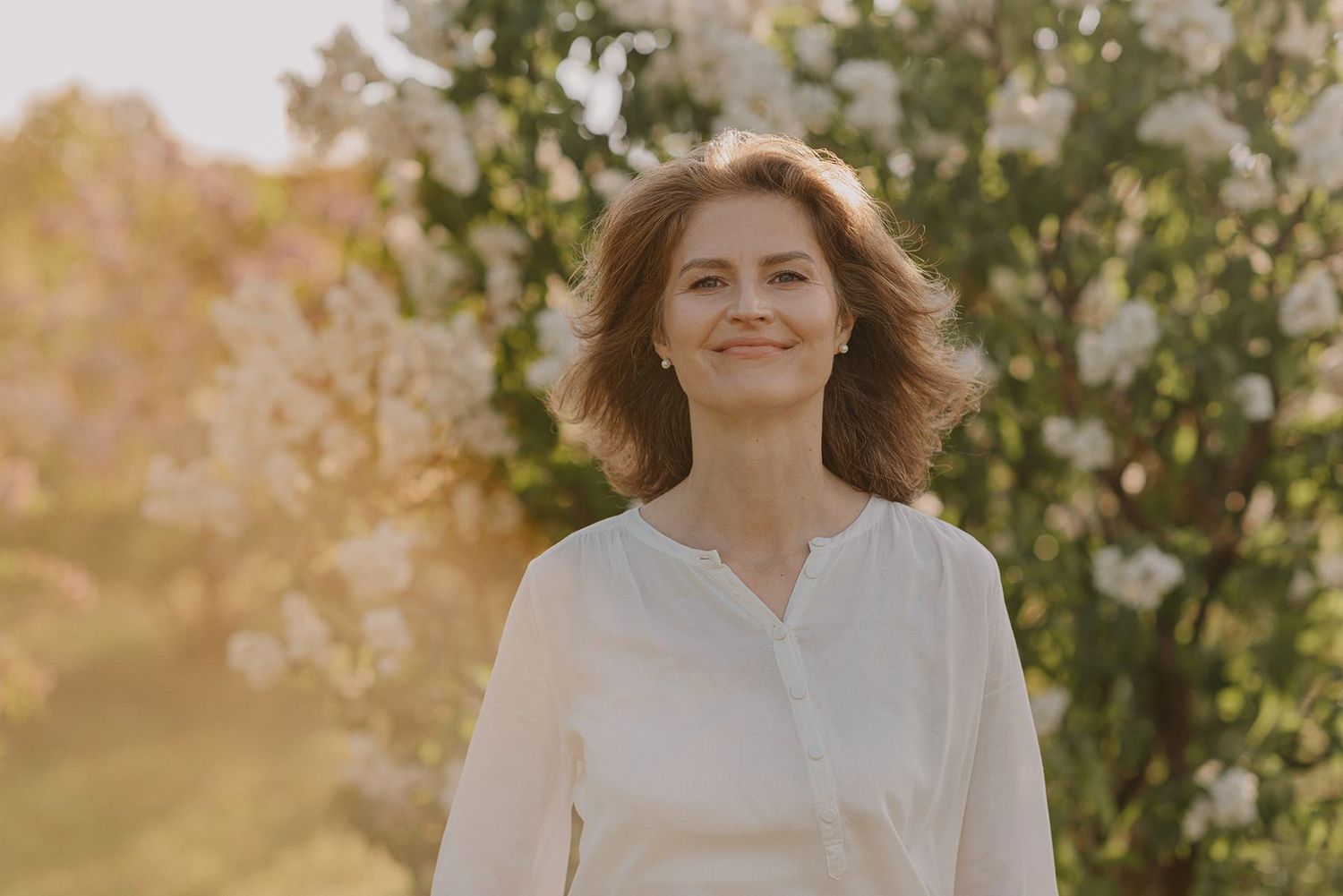 A person in a white blouse poses in warm sunlight against lush green foliage.