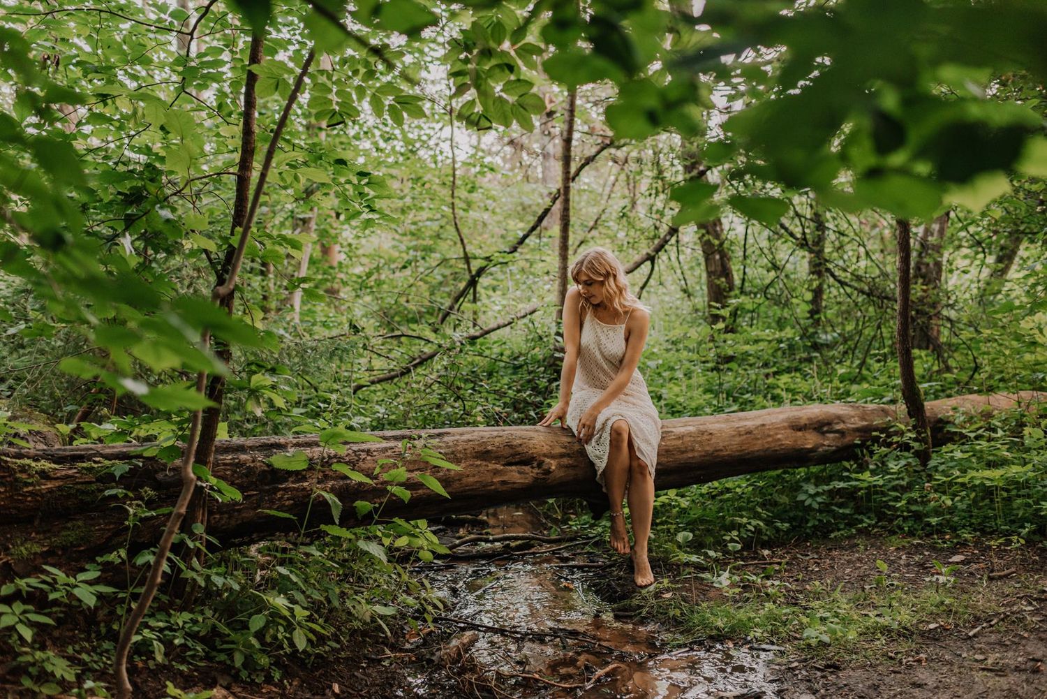 A woman in a white dress sits contemplatively on a fallen log in a lush forest.