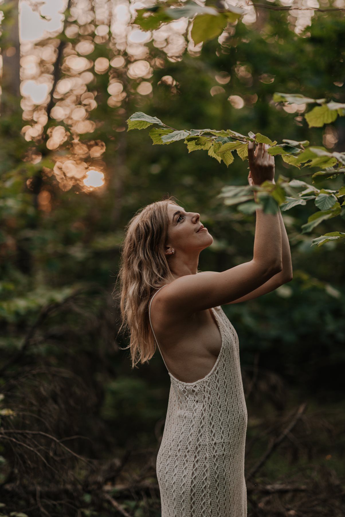 A woman in a knit dress reaches up to touch tree leaves in dappled sunlight.