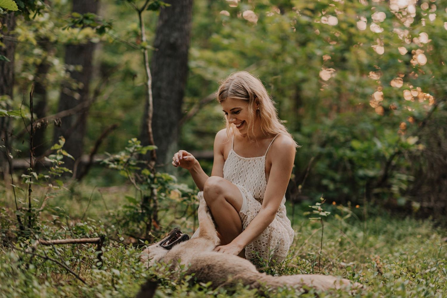 A woman in white lace dress sits gracefully on forest ground surrounded by greenery.
