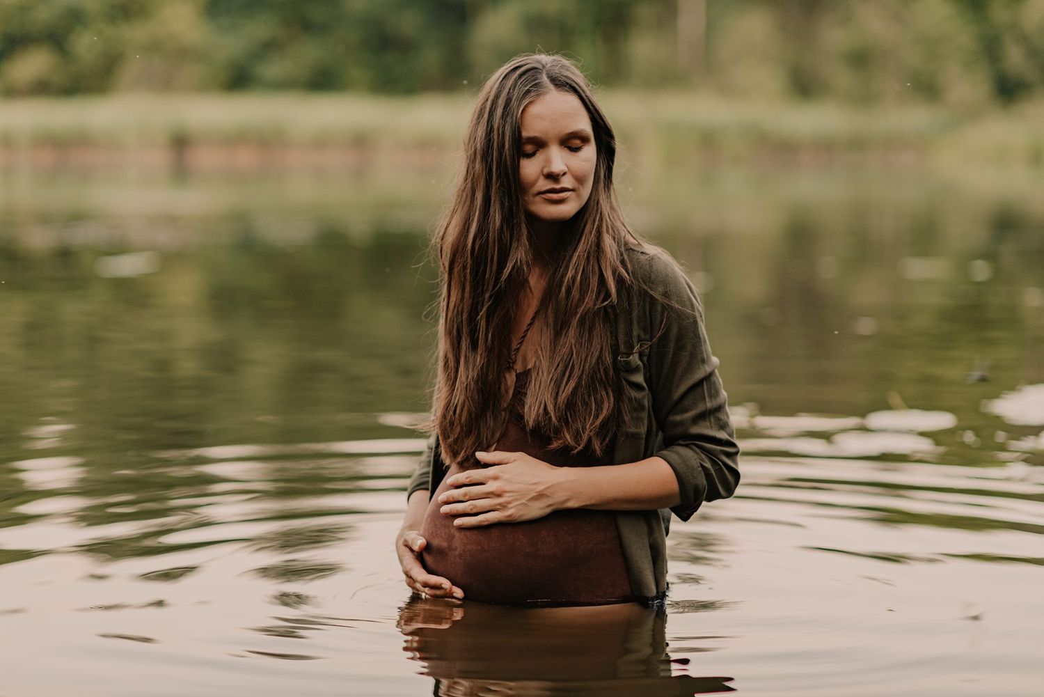 Person in a sheer dress stands in calm lake water during golden hour surrounded by forest scenery.