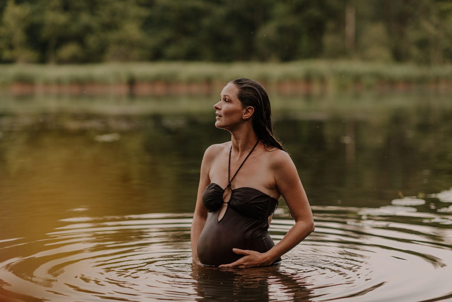 Figure wading in serene lake water with forest backdrop in artistic photoshoot.