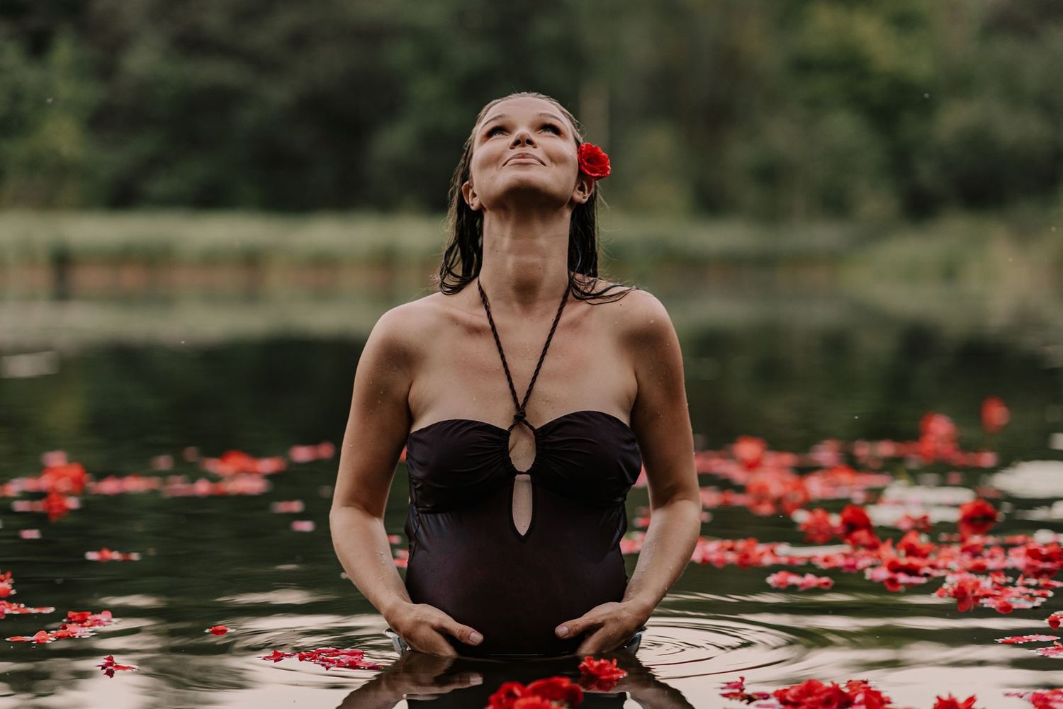Figure in dark dress poses dramatically in water with scattered red flowers.