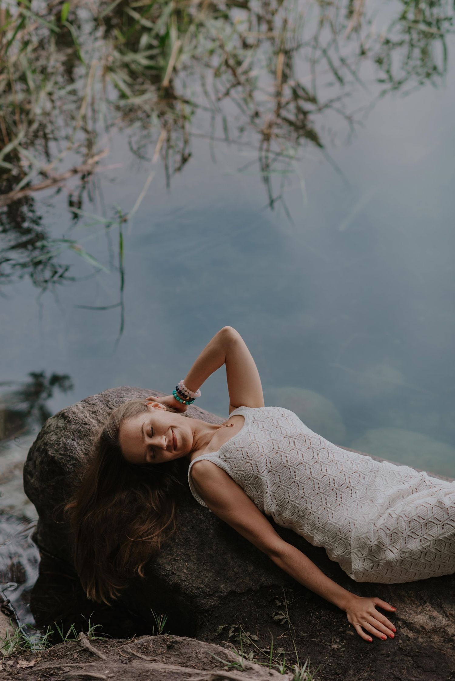 Dreamy portrait by a lake with someone reclining on rocks surrounded by hanging branches.