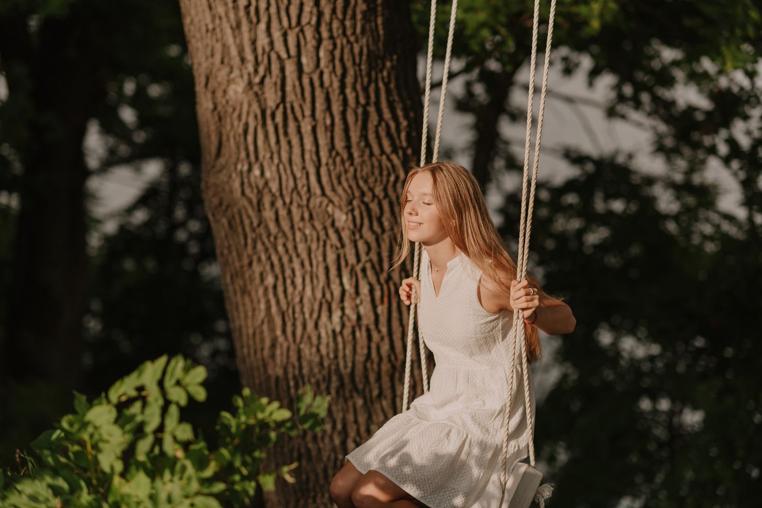 Person sitting on tree swing in white dress surrounded by greenery.