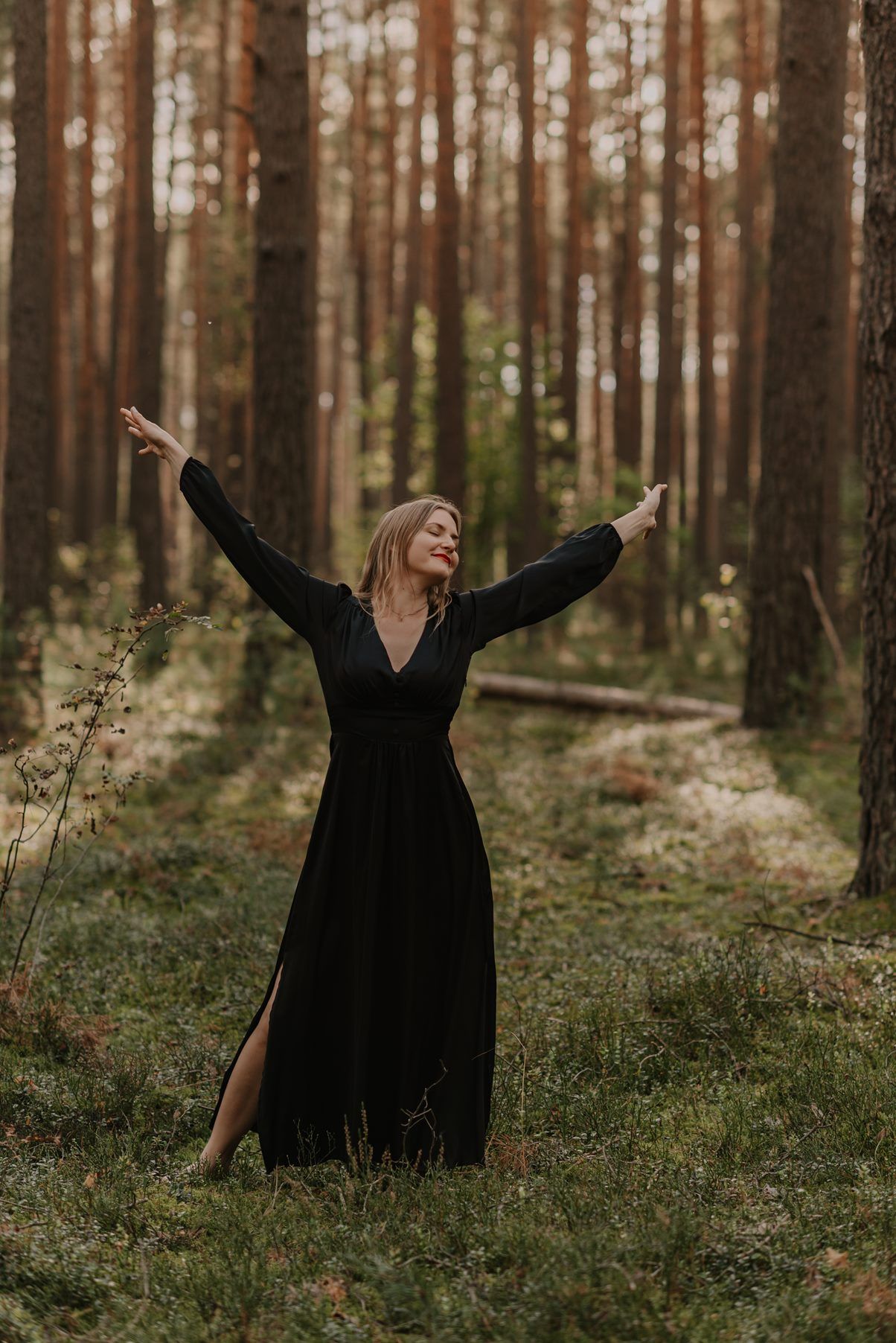A person in a black dress twirls joyfully among tall pine trees in a serene forest setting.