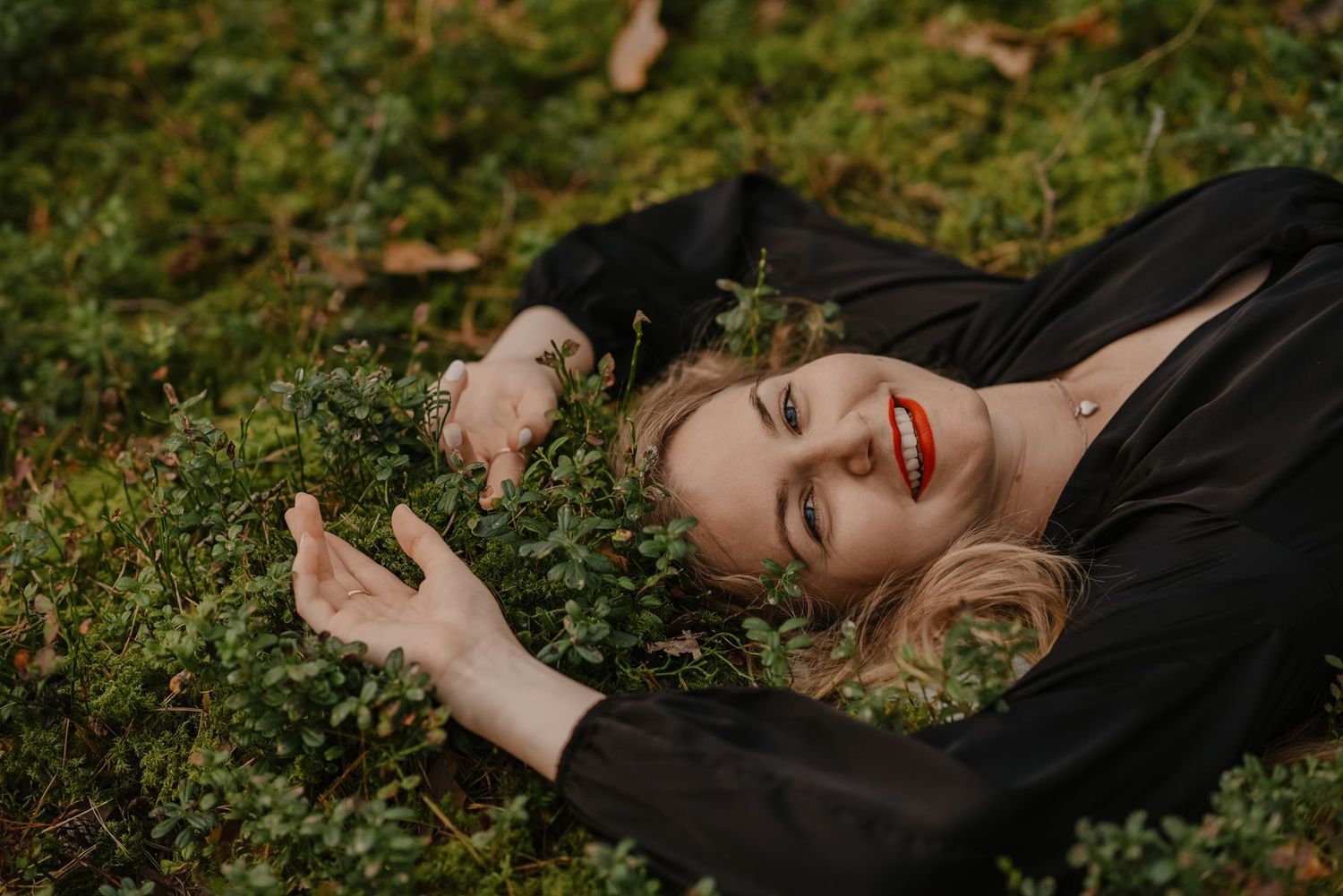 A figure in black lies on forest ground while smiling and posing expressively.