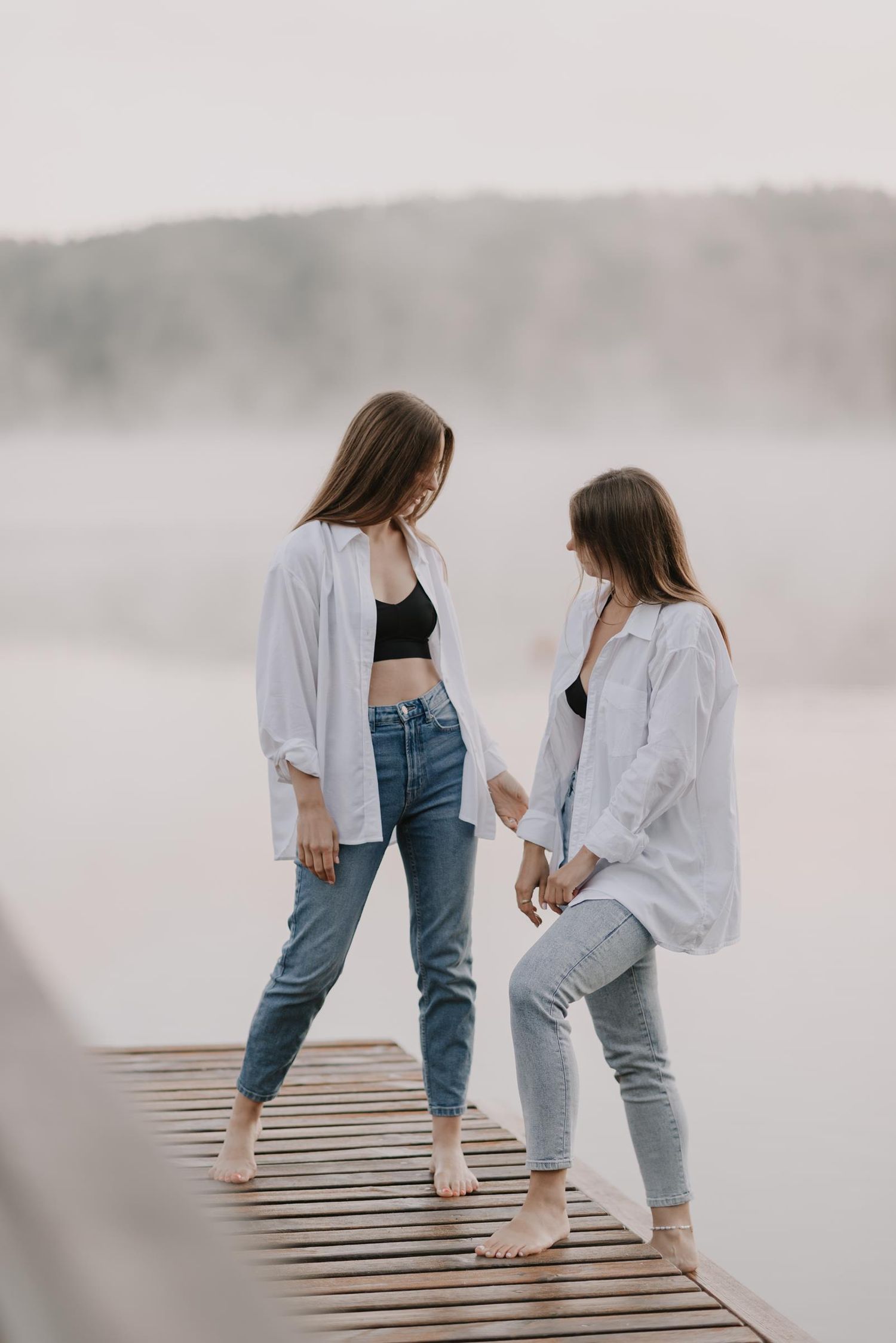 Two people in casual outfits stand barefoot on a misty wooden dock.