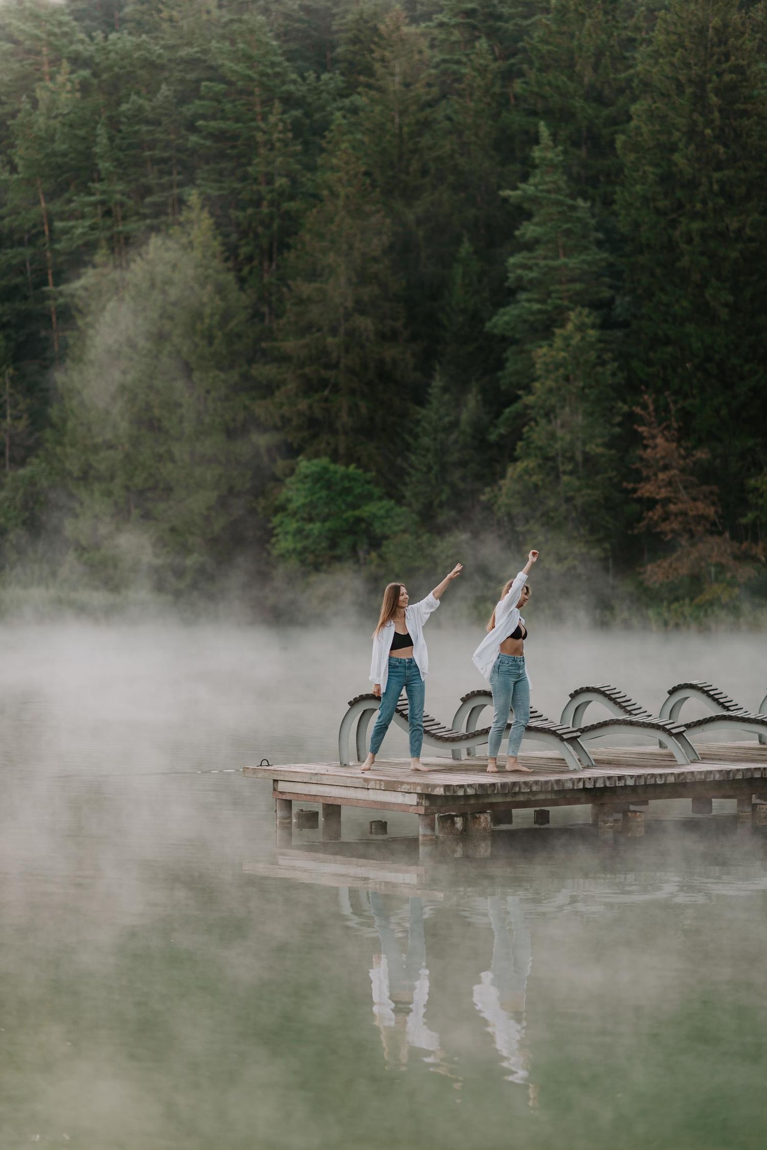 Two figures in denim stand with raised arms on a dock surrounded by morning mist and evergreen trees.