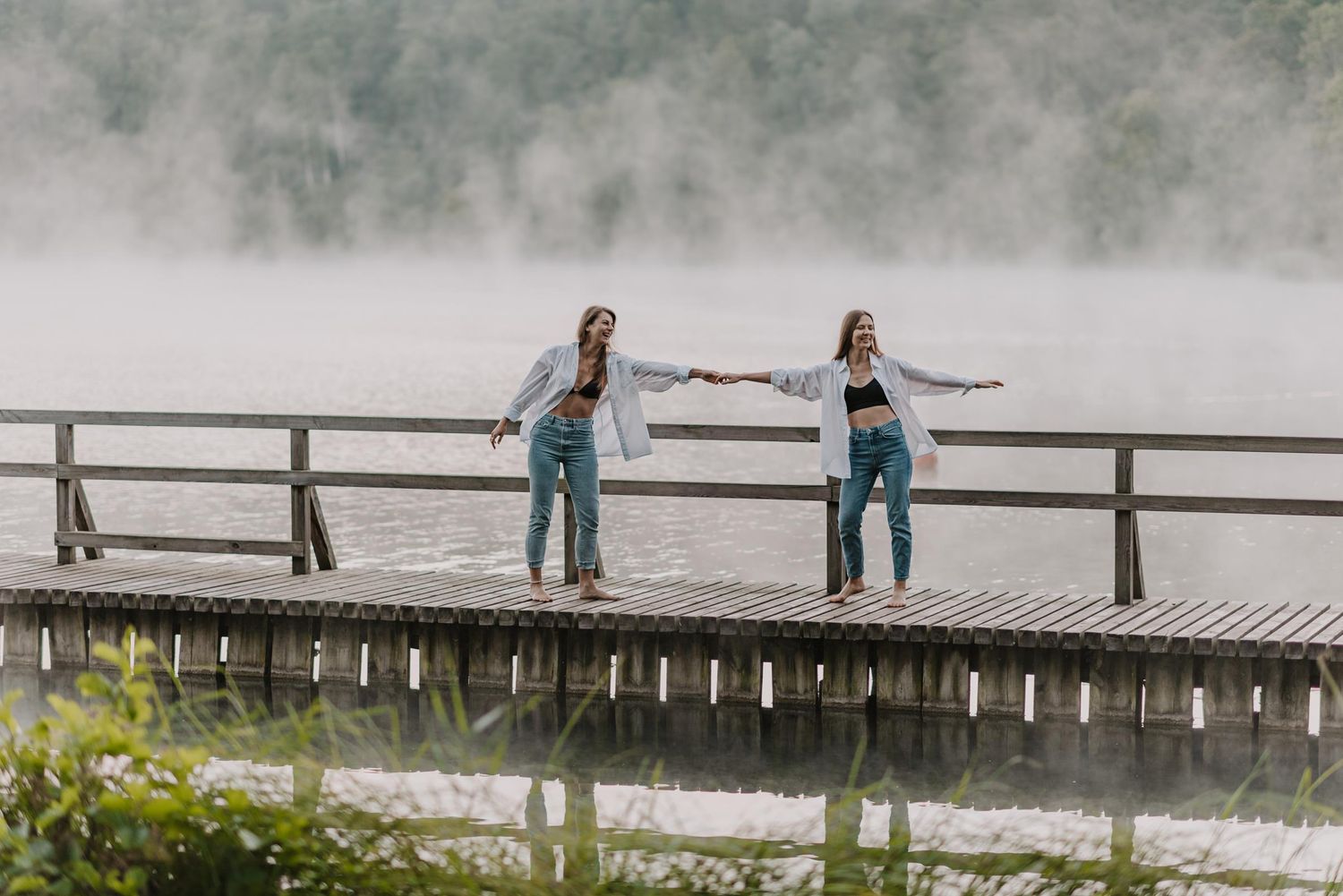 Two people pose playfully on a wooden bridge over misty waters.
