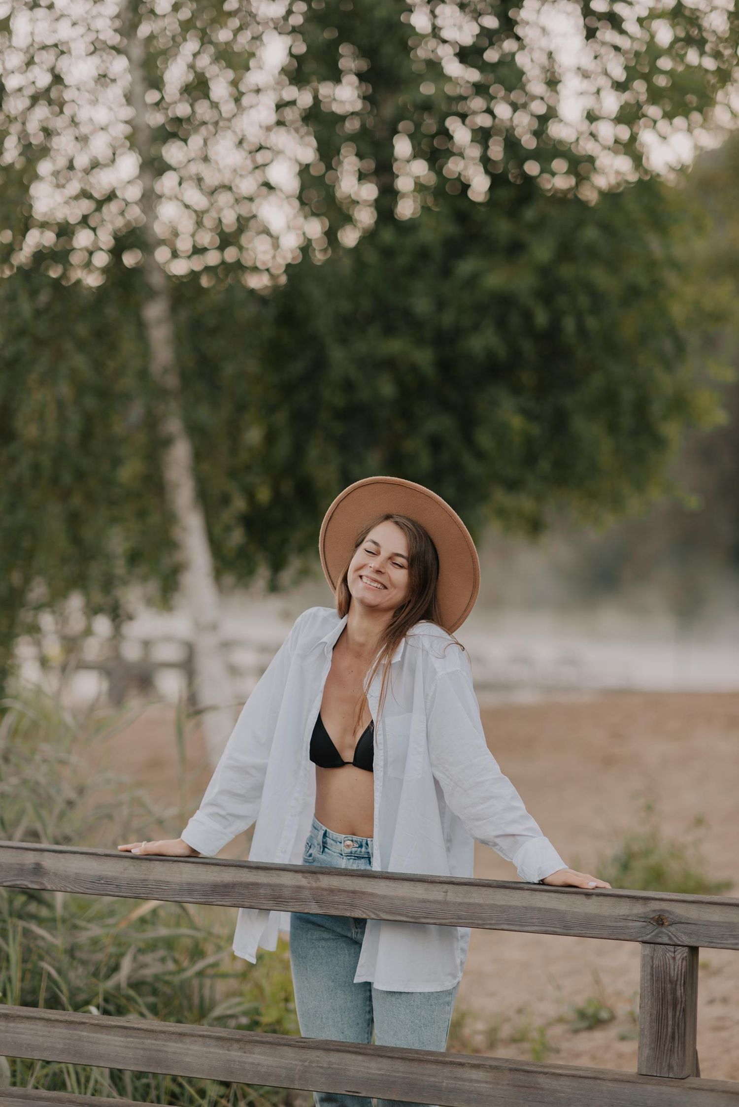 Person in brown hat and denim leans against wooden railing in dreamy summer setting.