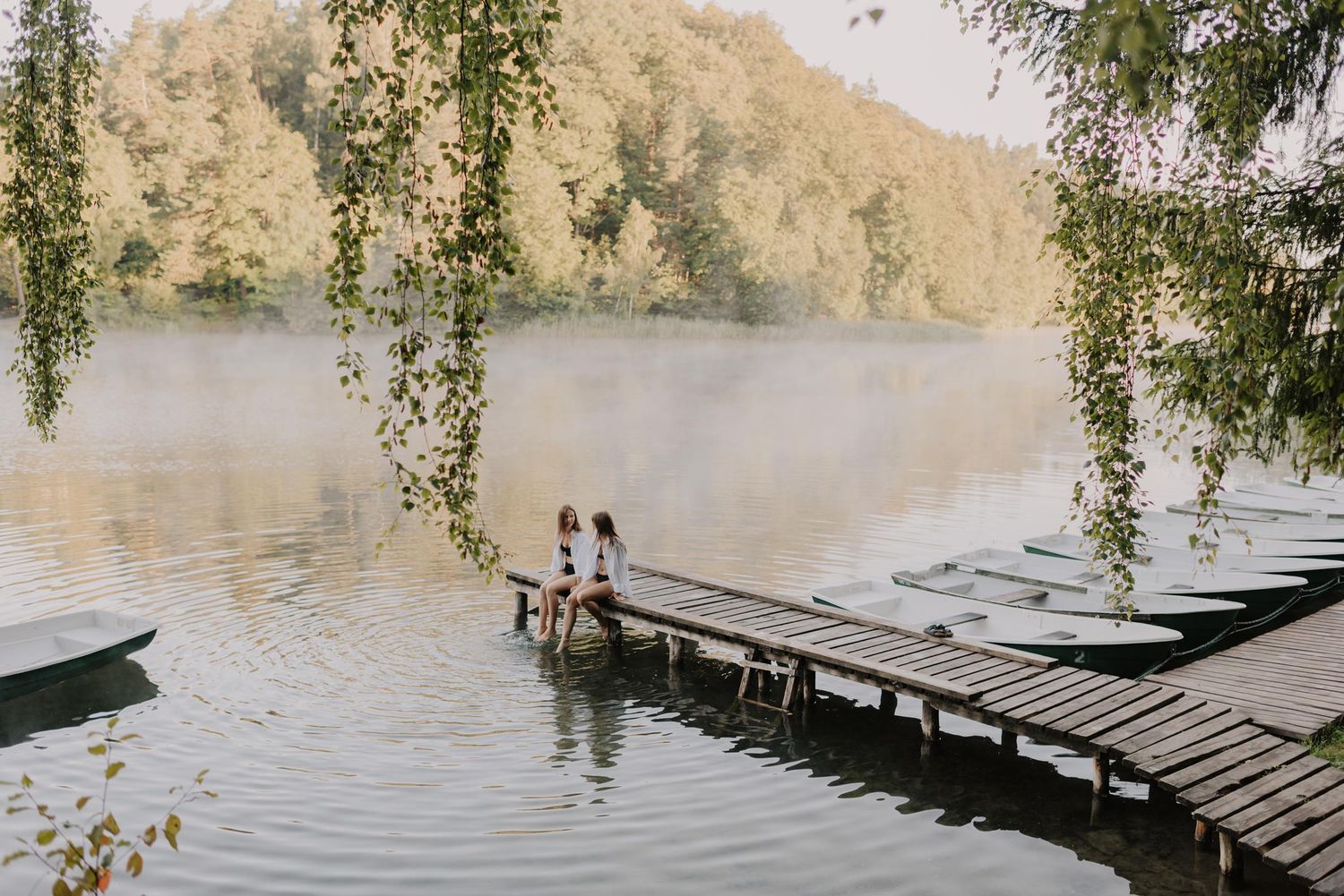 Two silhouettes stand on wooden dock with willow trees and calm lake in background.