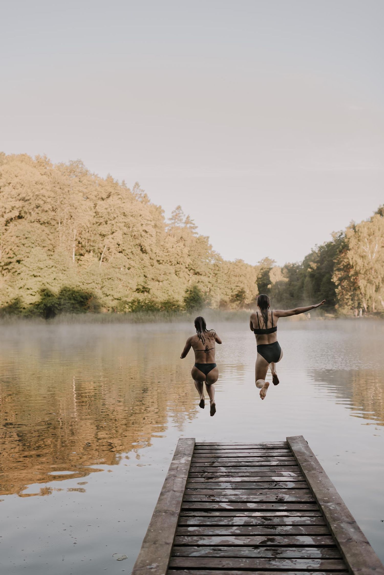 Two people leap joyfully off wooden dock into peaceful lake at sunset.