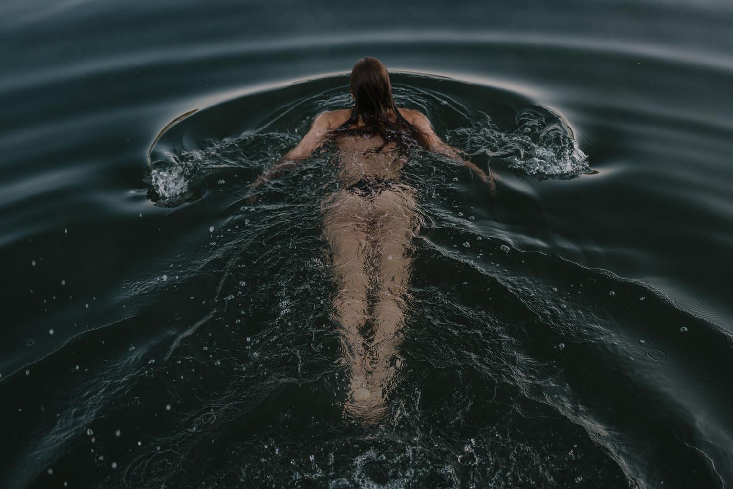 Person swimming in dark waters creates ripples while floating on their back.